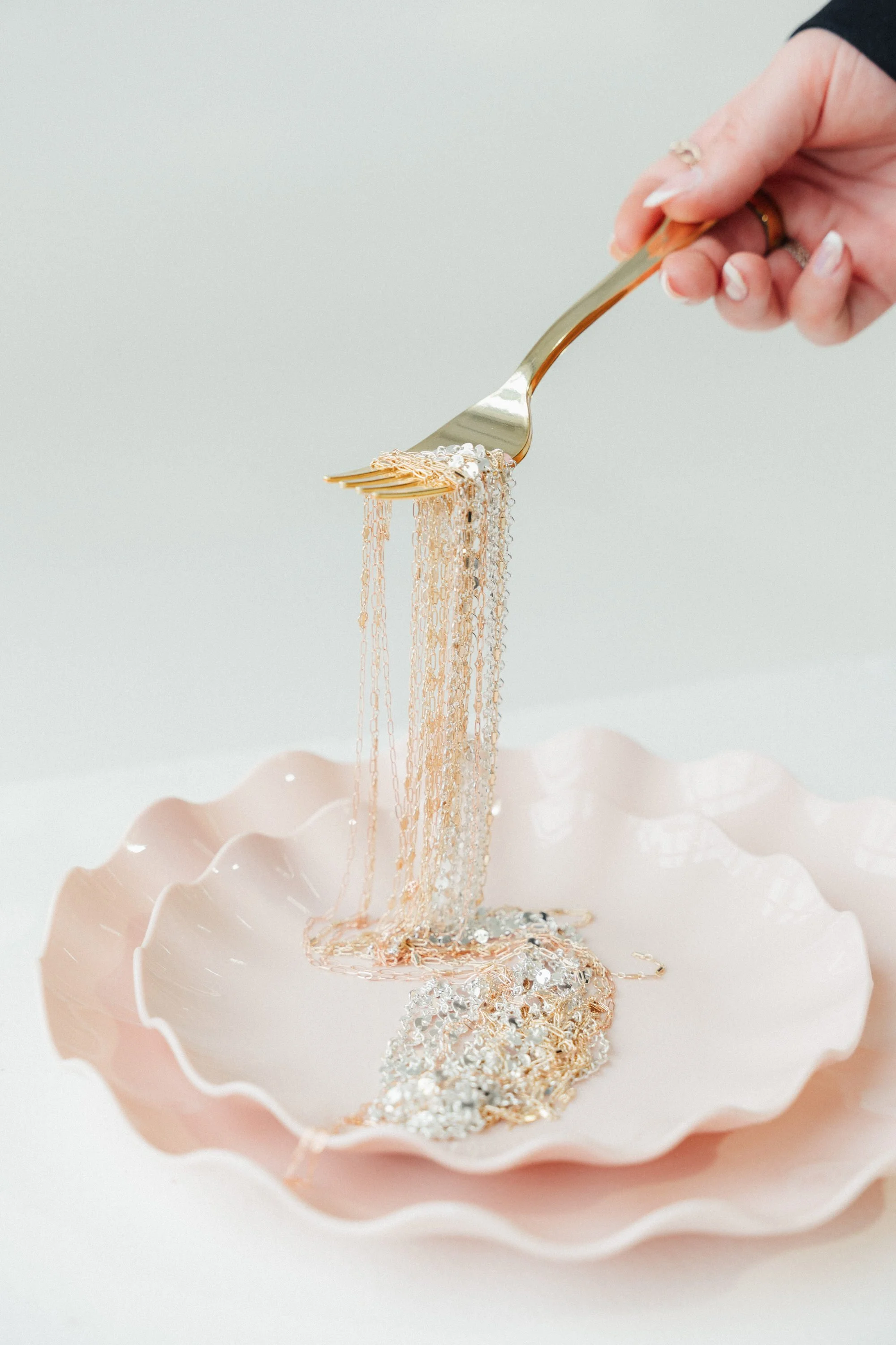 A hand holding a gold fork with shiny silver chains and jewelry spilling onto a pink ruffled bowl.