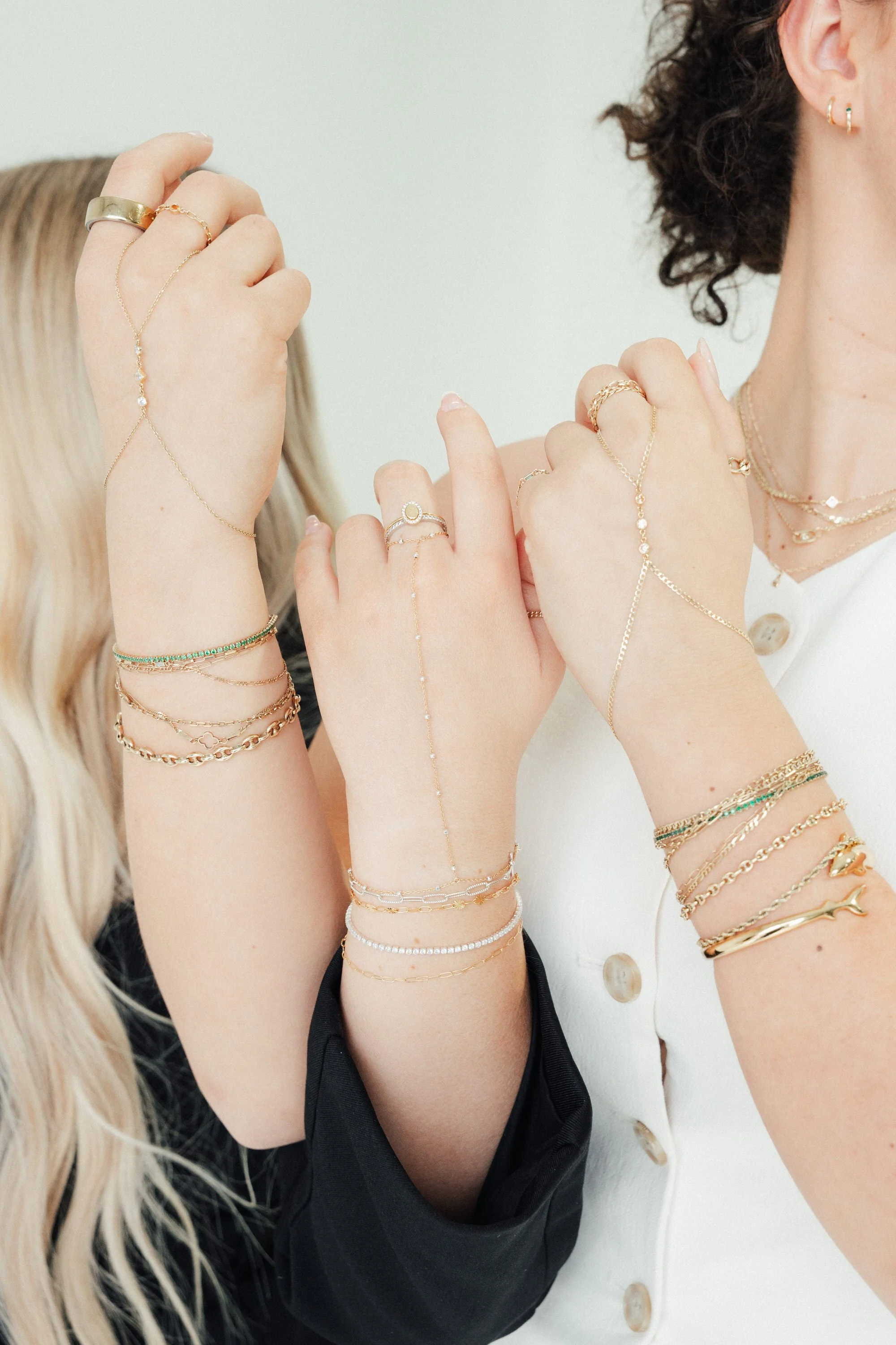 Close-up of two women showing their arms with numerous gold and silver jewelry, including rings, bracelets, and layered necklaces.