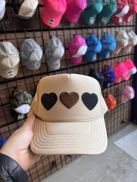 A beige baseball cap with three heart patches in black, brown, and navy on the front, held by a person in front of a display of colorful caps on a grid wall.