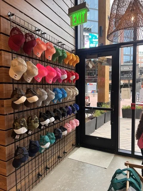 Colorful hats and caps displayed on a black wire grid wall in a retail store, next to a glass door with an open green exit sign above.