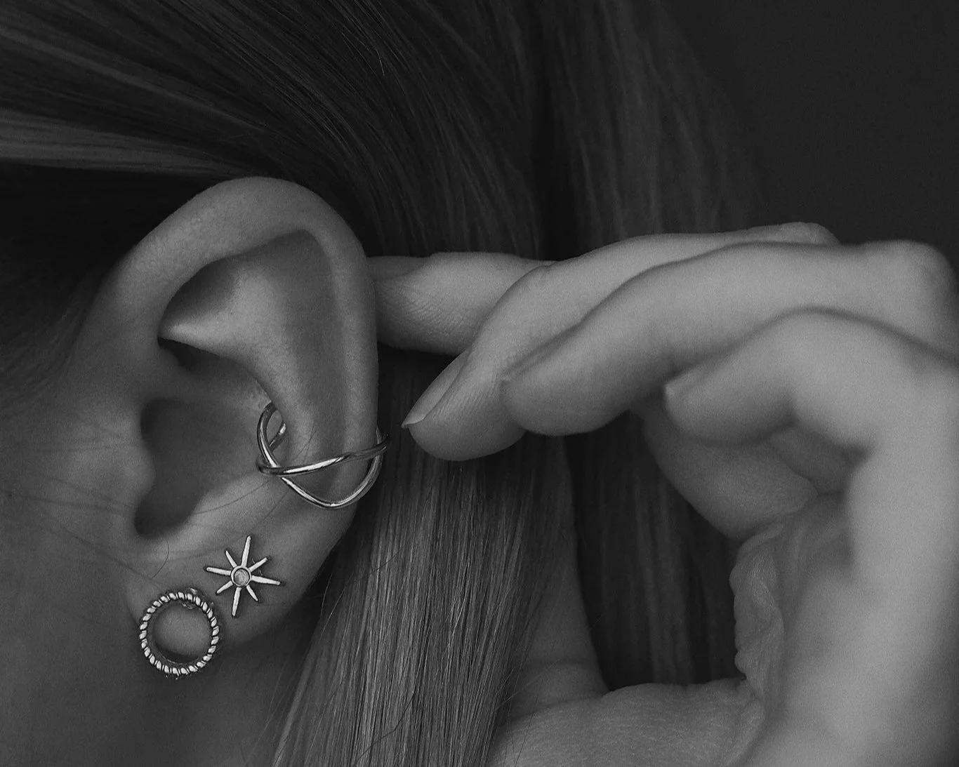 Close-up of a person’s ear adorned with three earrings: a small hoop in the lobe, a sun-shaped stud, and a beaded hoop. The person is holding their earlobe with one hand, and their hair is visible in the background.