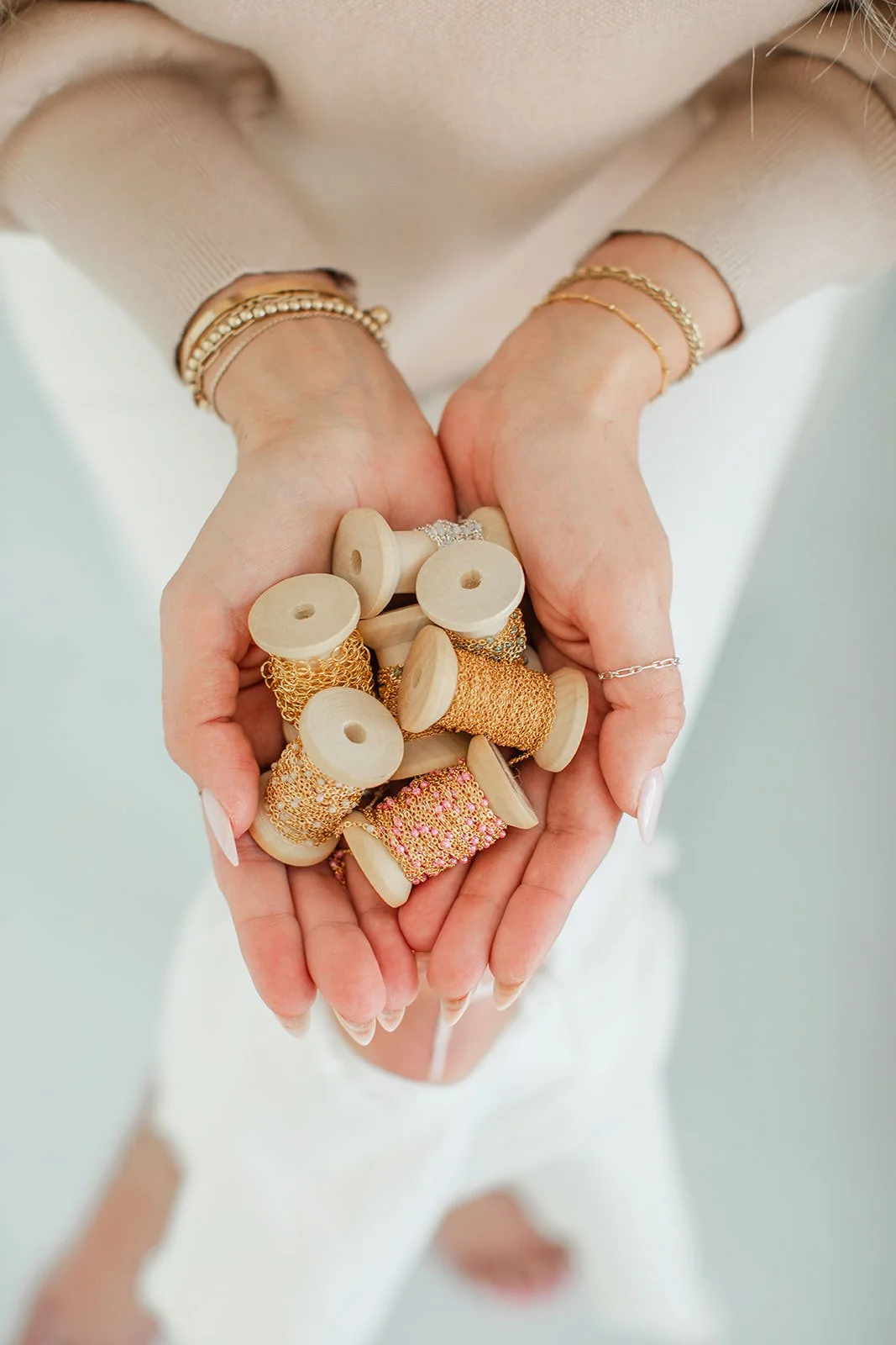 A person holding a handful of wooden spools with gold and pink thread. The person is wearing multiple gold bracelets and a silver ring, and has a beige sweater. The background is minimal and light-colored.