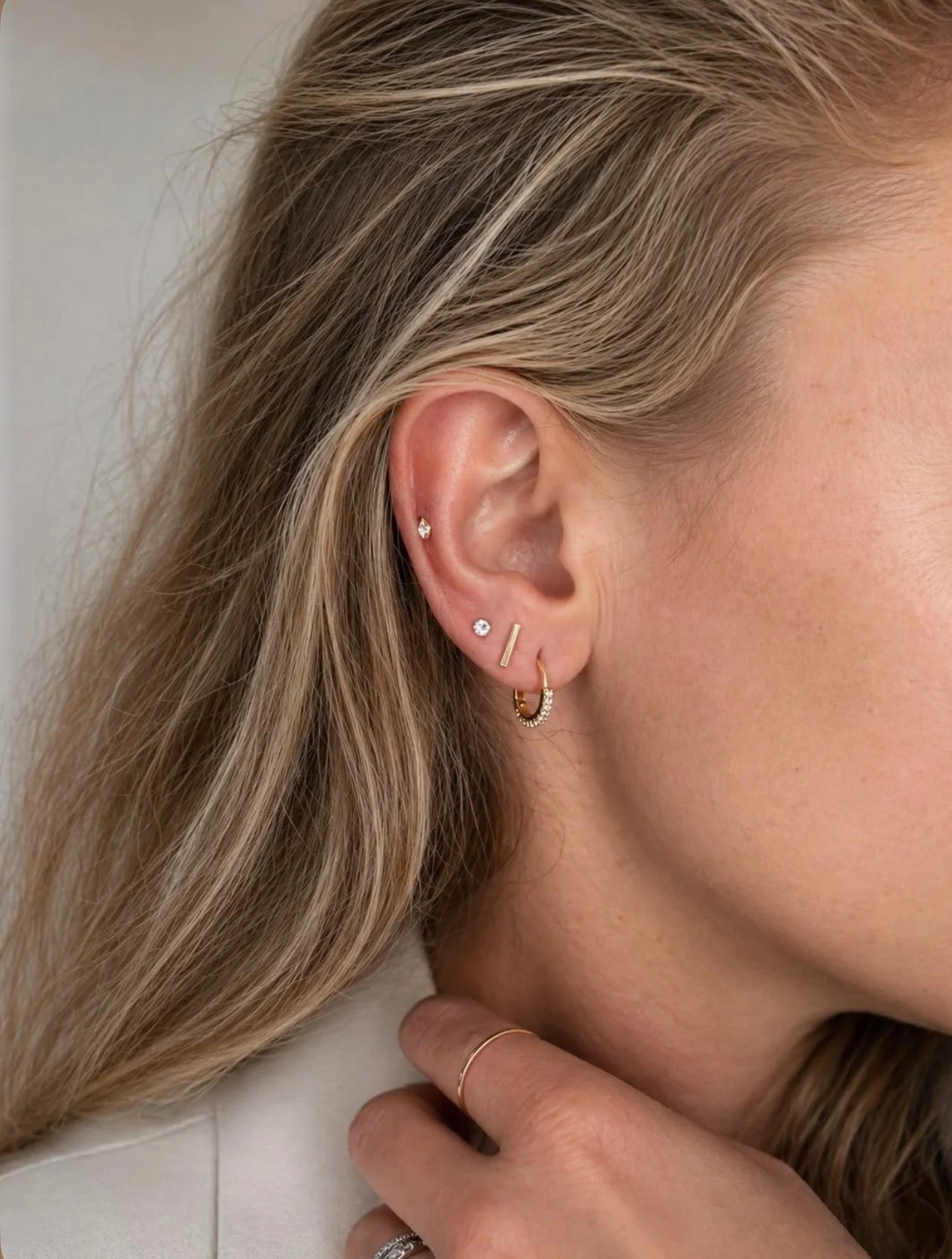 Close-up of a woman's ear with multiple earrings, including a small hoop with diamonds, a vertical bar earring, a stud with a gemstone, and a small diamond stud. The woman's blonde hair partially covers her ear and face, and she wears a thin ring on her finger.