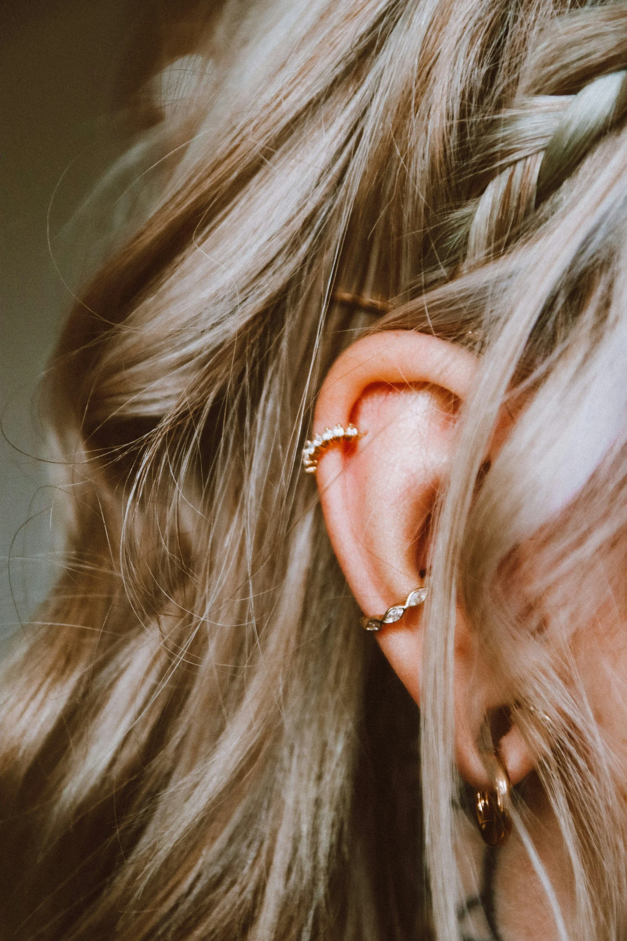 Close-up of a woman's ear adorned with multiple earrings, including a curved diamond and gold ear cuff, with long blond hair covering part of the ear.