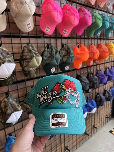 A turquoise trucker cap with embroidered patches, held in front of a display of colorful caps on a wire grid wall.