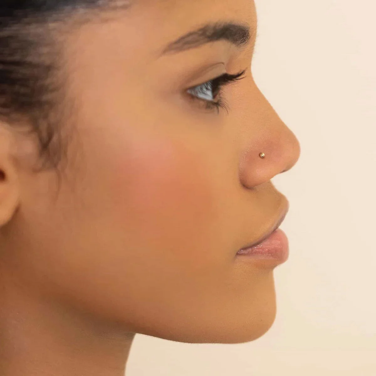 Side profile of a woman with a small nose piercing, smooth skin, and subtle makeup.