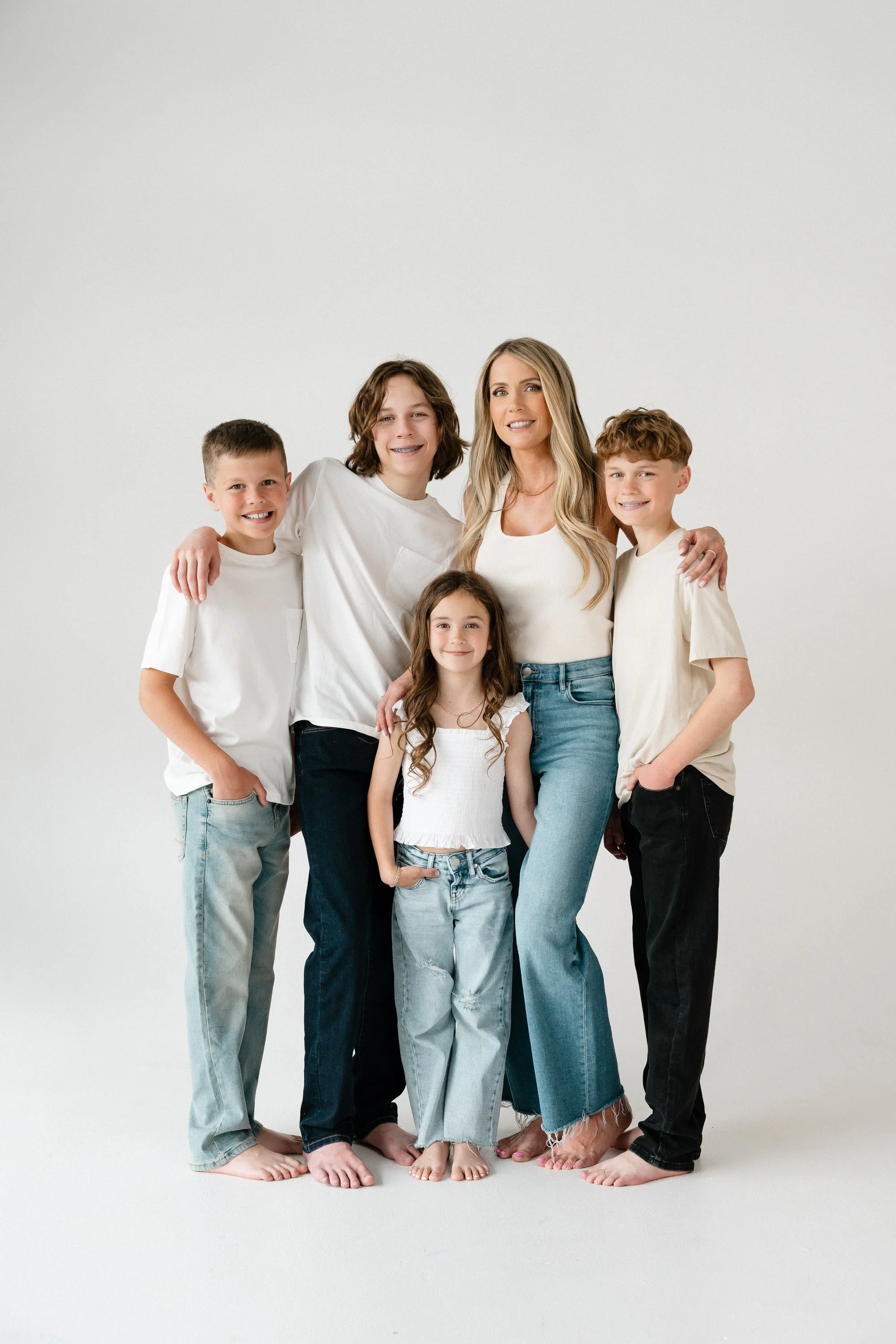 A woman and five children, all in casual white or light-colored clothing, standing together barefoot against a plain white background, smiling and embracing each other.