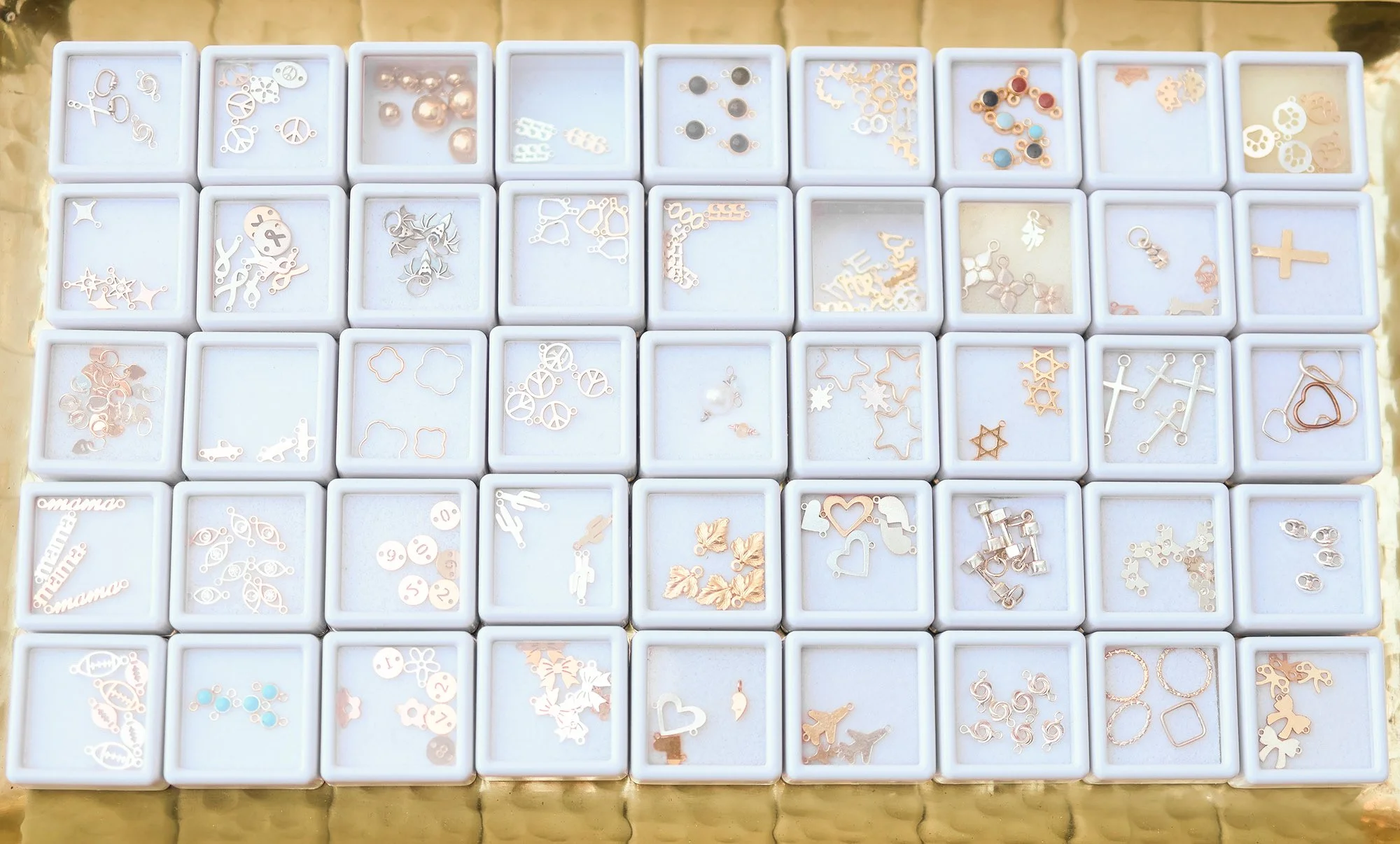 A display of small jewelry charms in white boxes, including crosses, hearts, stars, animals, and abstract shapes, arranged in rows.