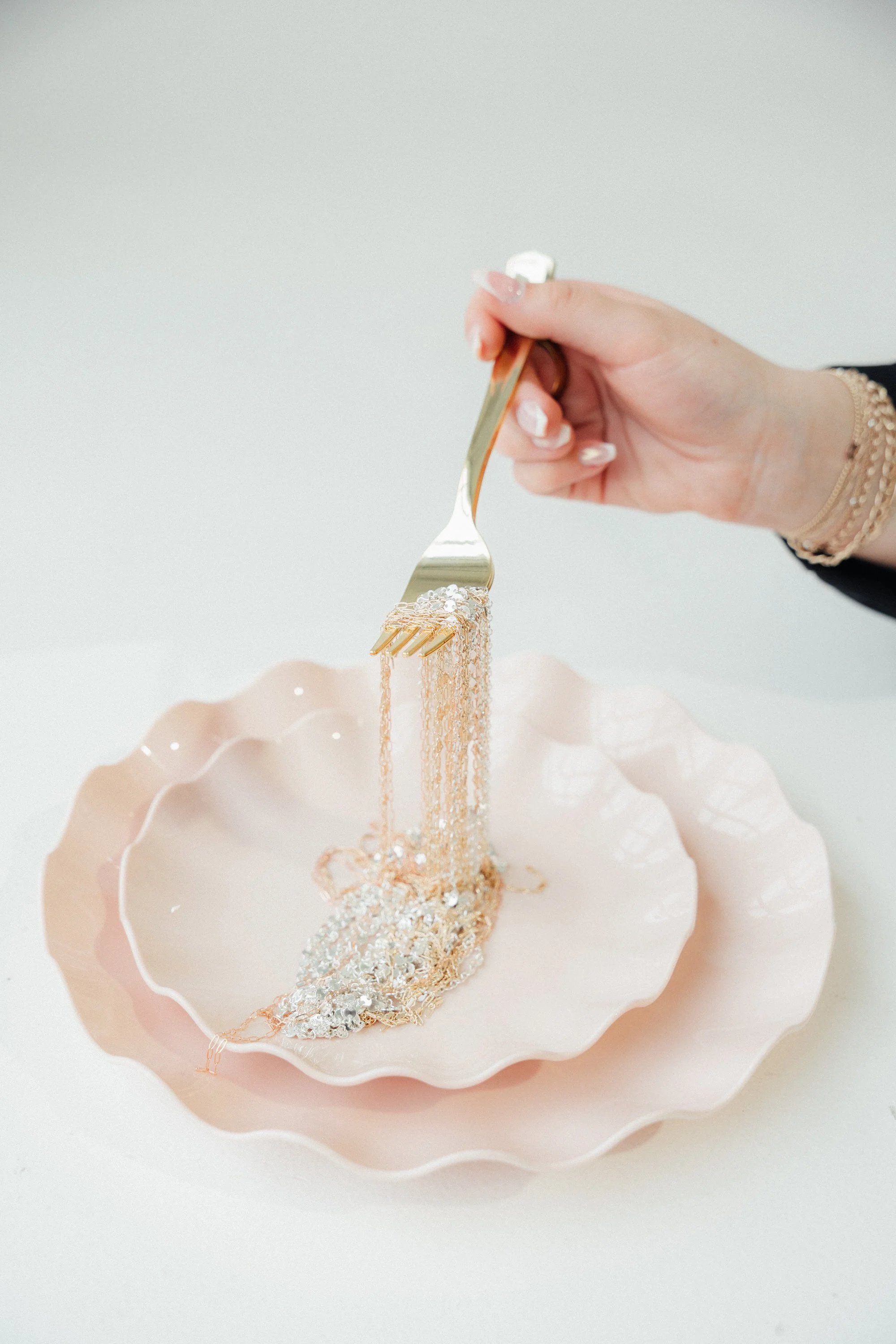 A hand is holding a gold-colored fork with a piece of jewelry on it over a pink fluted bowl filled with silver glitter and decorative beads, with another similar bowl in front.