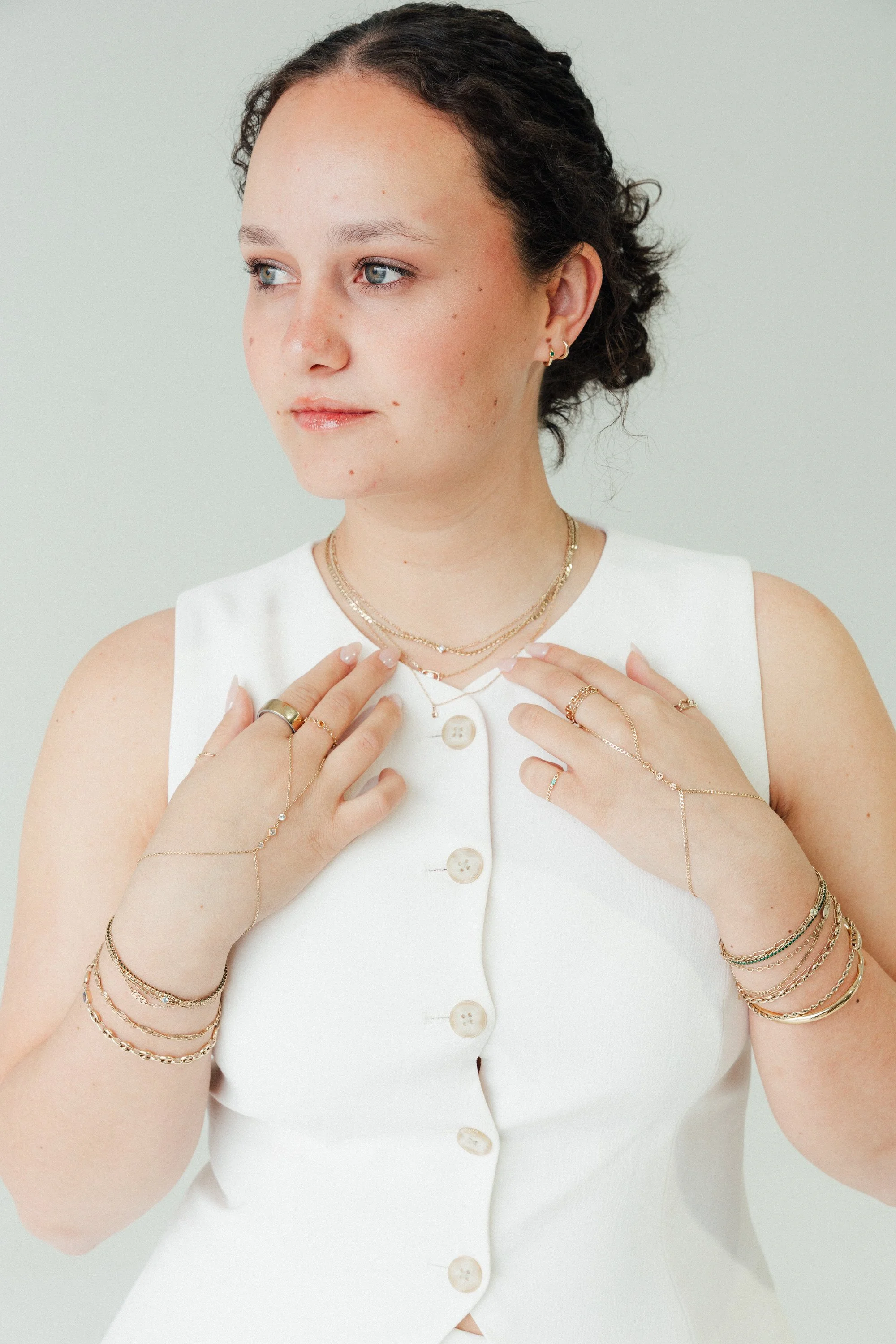 A woman with curly brown hair pulled back, wearing a sleeveless white dress with buttons, multiple gold and silver jewelry, including rings, bracelets, necklaces, and earrings, and looking to her left against a plain light green background.