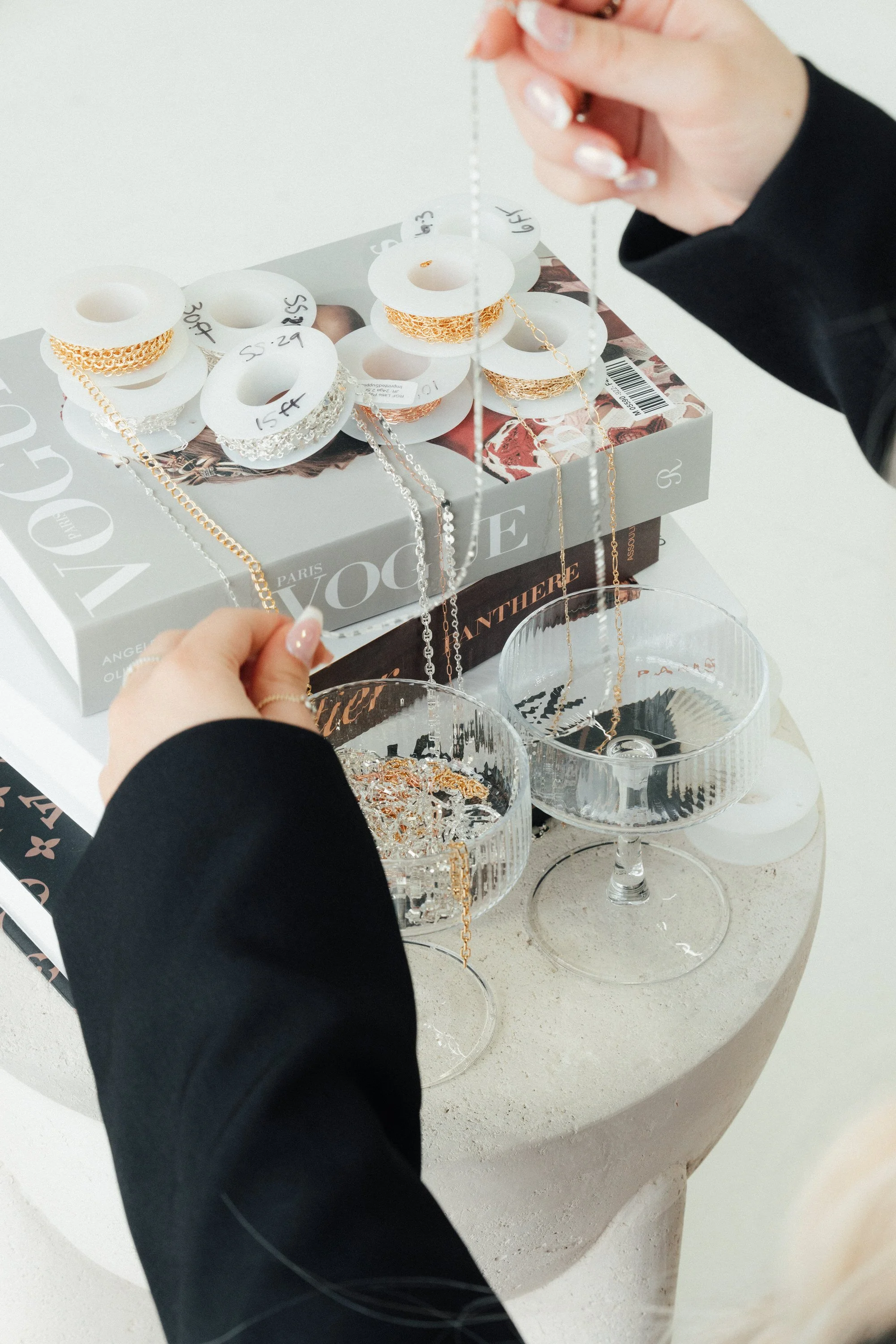 Person wearing black sleeve selecting gold and silver chain jewelry from display on a table with jewelry boxes and glasses.