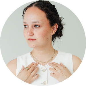 A young woman with curly hair wearing a sleeveless white top and layered necklaces, holding her hands near her chest.
