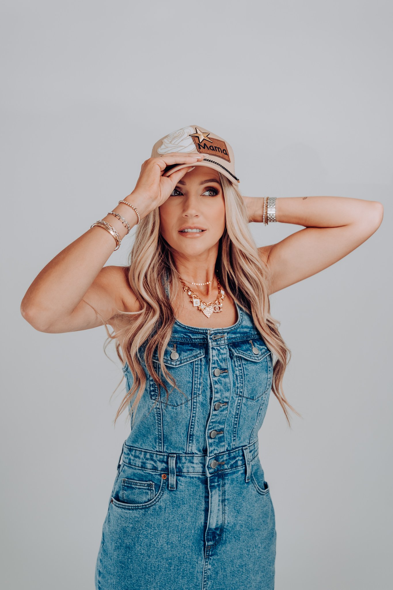Woman with long blonde hair wearing a denim dress and jewelry, holding a beige baseball cap with decorative patches reading 'Mama' against a plain gray background.
