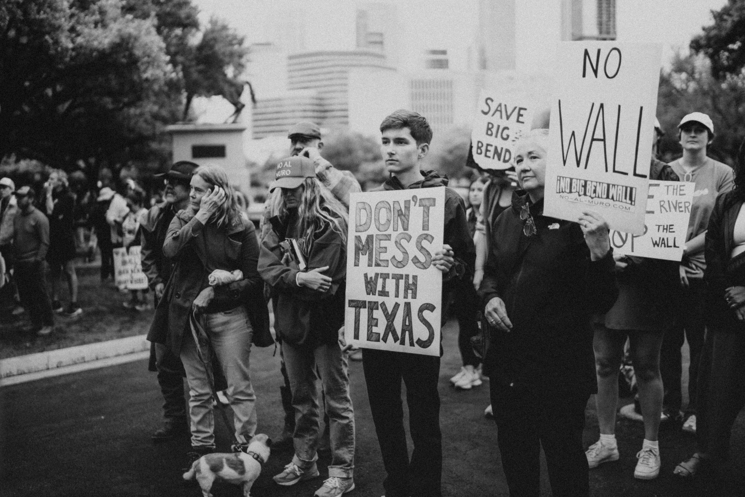 A group of people participating in a protest or rally, holding signs and banners advocating against the construction of the wall, with city buildings in the background.