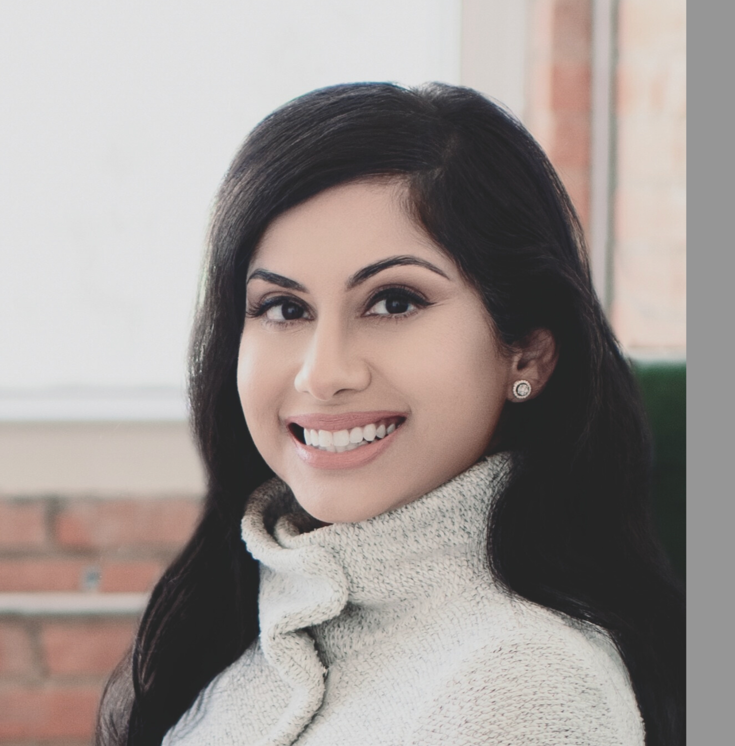 Portrait of a woman smiling, with dark hair, wearing earrings and a turtleneck sweater, sitting indoors with brick and window background.