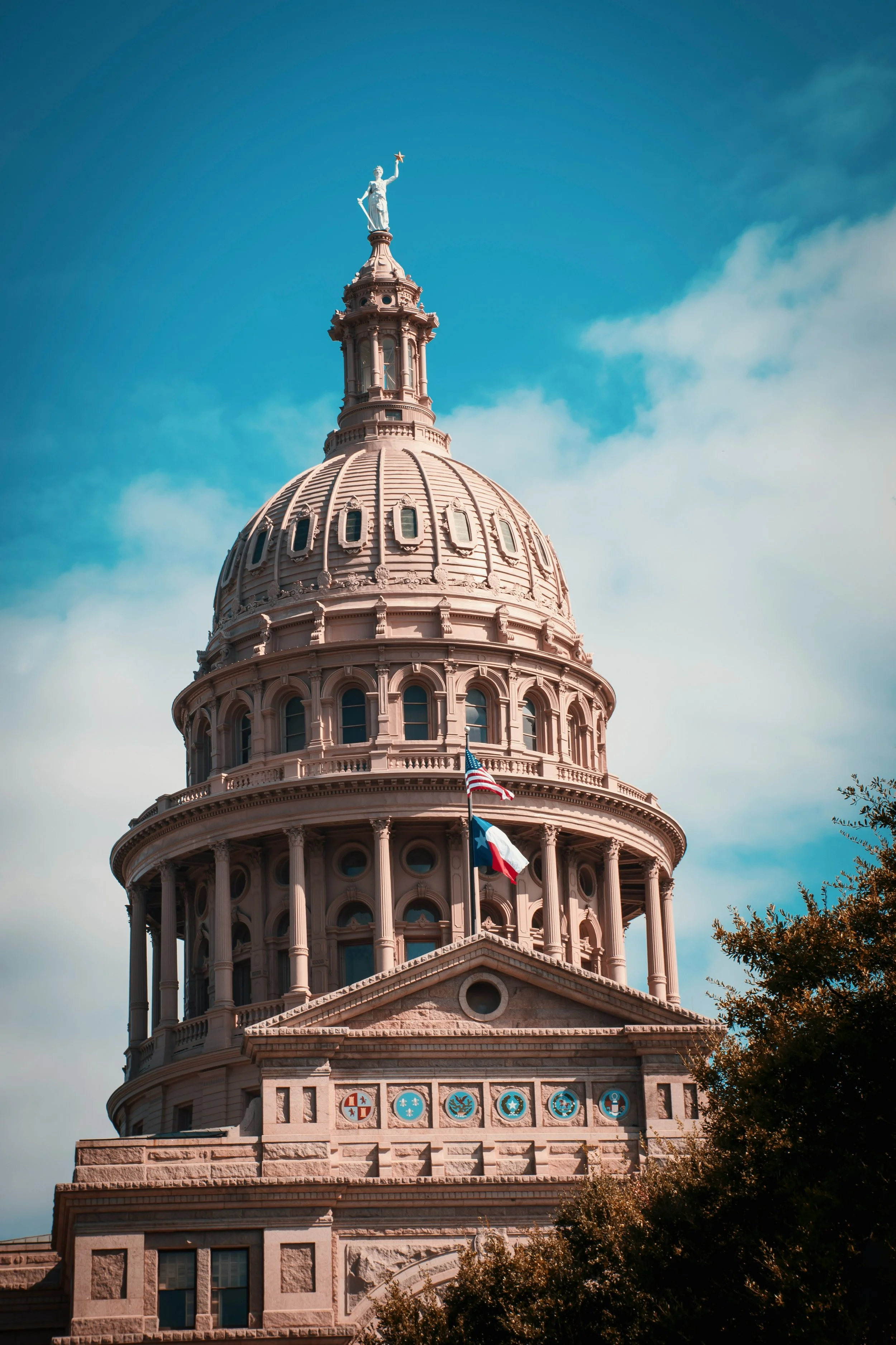 Close-up of the Texas State Capitol building dome with a statue on top, flags, and decorative symbols below, under a partly cloudy sky.
