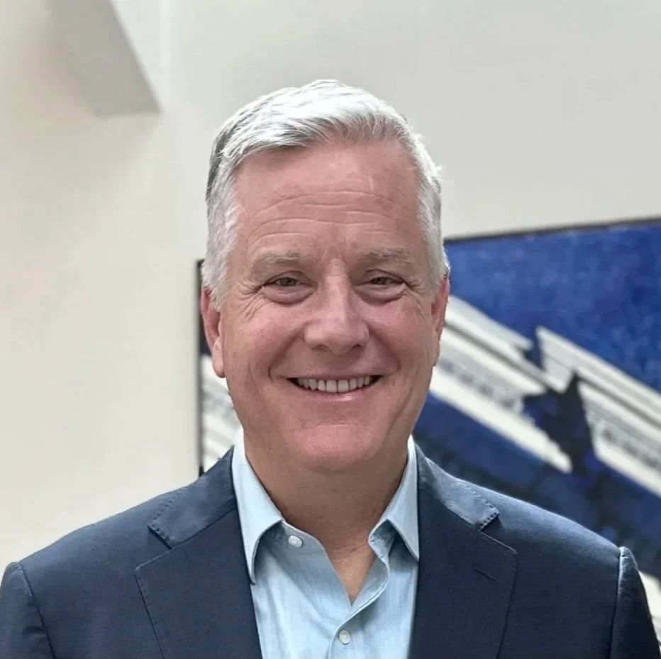 Smiling middle-aged man with gray hair in a suit and light blue shirt, standing indoors near a staircase with blue carpeting.