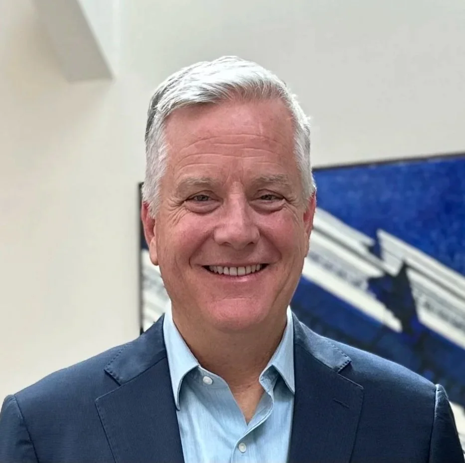 Portrait of a smiling middle-aged man with gray hair, wearing a dark suit jacket and light blue collared shirt, in an office setting.