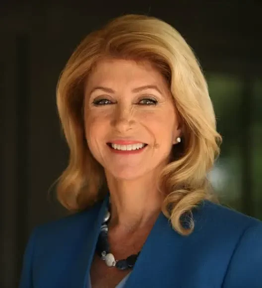 Portrait of a middle-aged woman with blonde hair, smiling, wearing a blue blazer and pearl earrings, in an indoor setting.