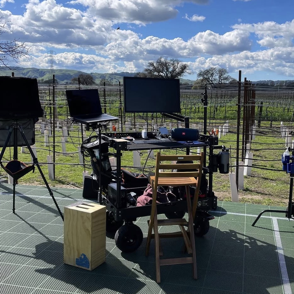 A camera setup on a mobile cart with multiple monitors, cables, and electronic equipment set on a platform outdoors in a vineyard on a sunny day with clouds. Andrew Fornasier Digital Tech San Francisco