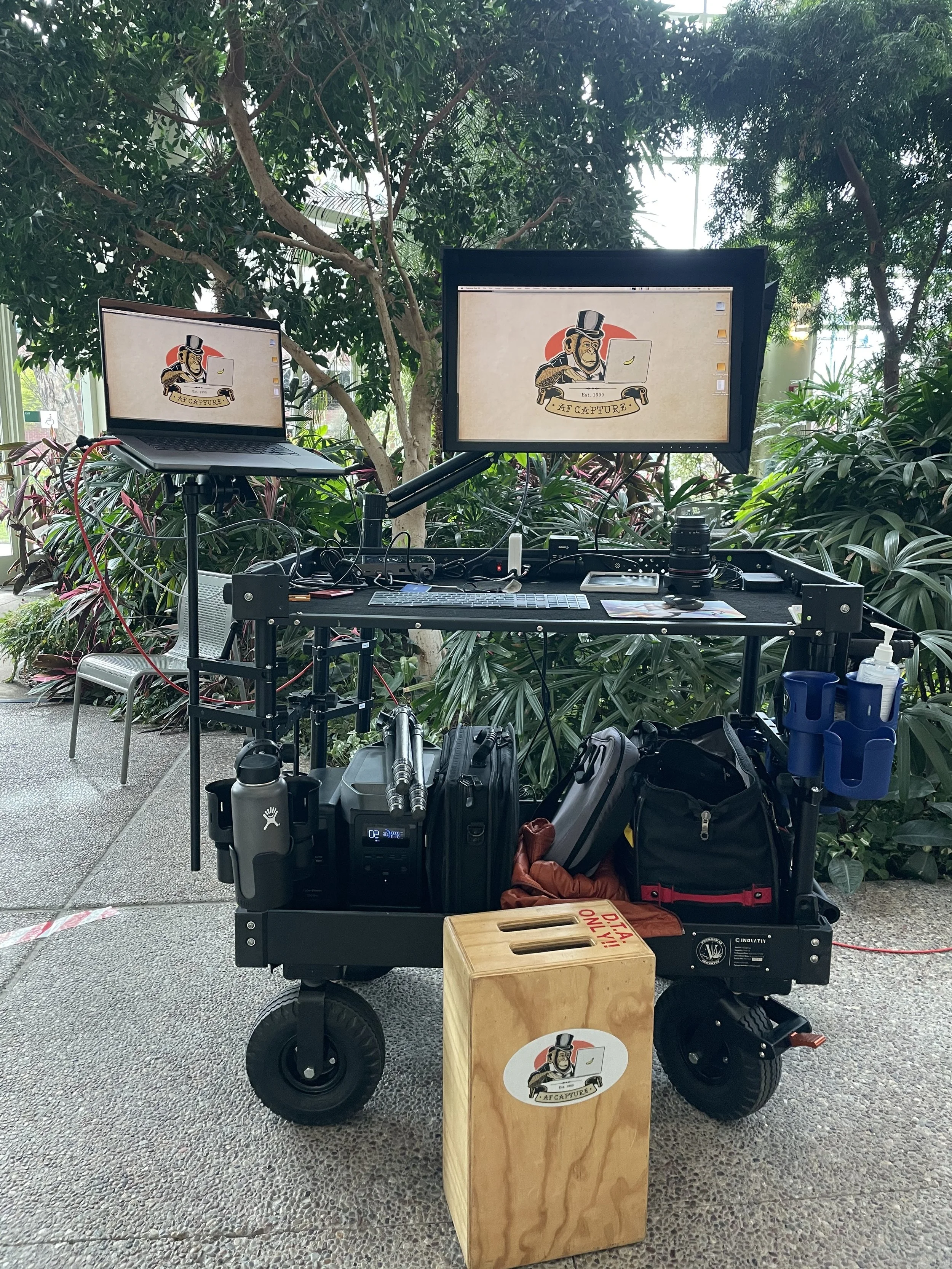 A portable photography setup with a table holding camera gear and two monitors displaying a logo of a cartoon gorilla wearing a top hat with the text "AF Capture". The setup is outdoors among lush green plants and trees. Andrew Fornasier Digital Tech