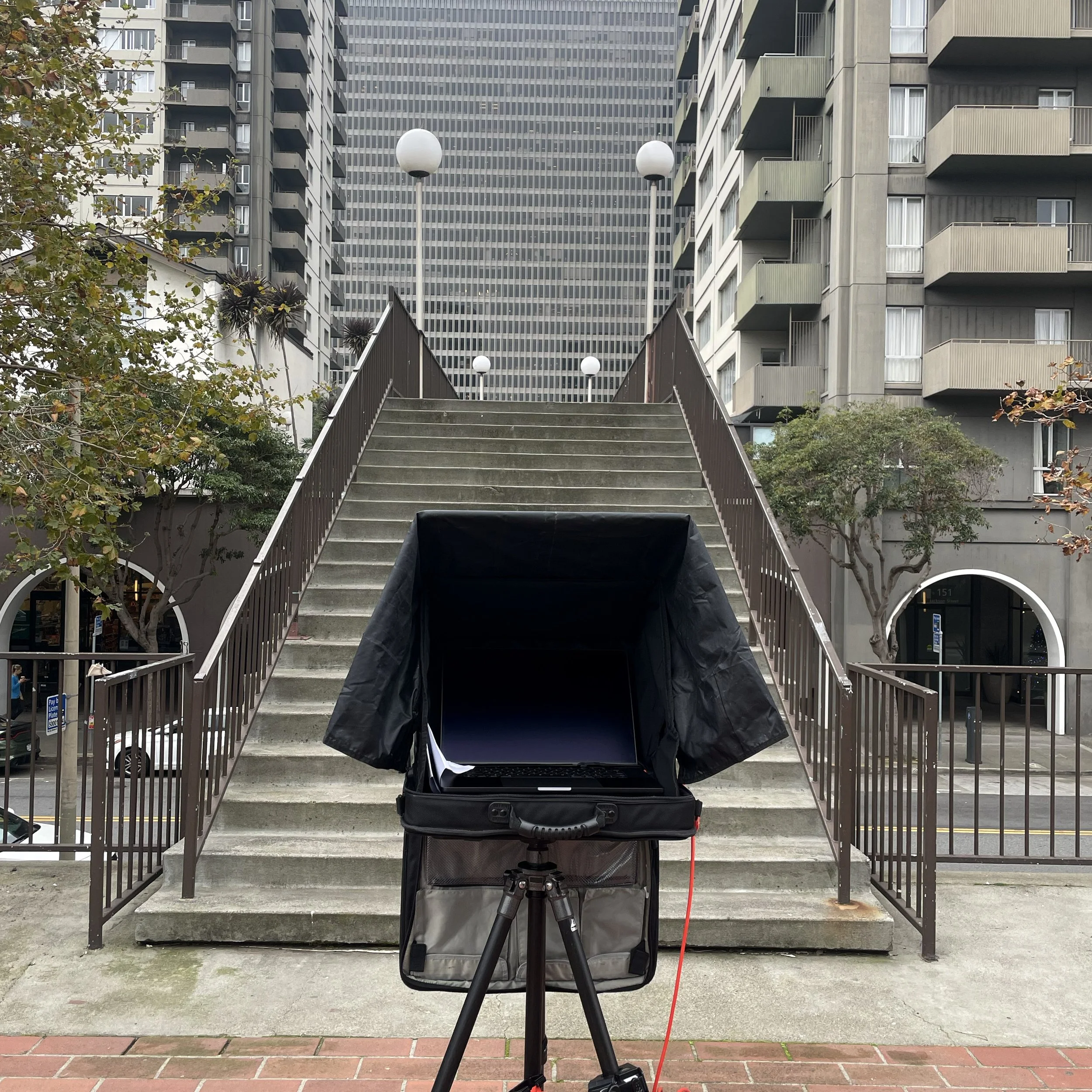 Urban staircase with railing on both sides, leading up through a cityscape with high-rise buildings, trees, and street lamps, with a laptop on a tripod standing at the bottom of the stairs.