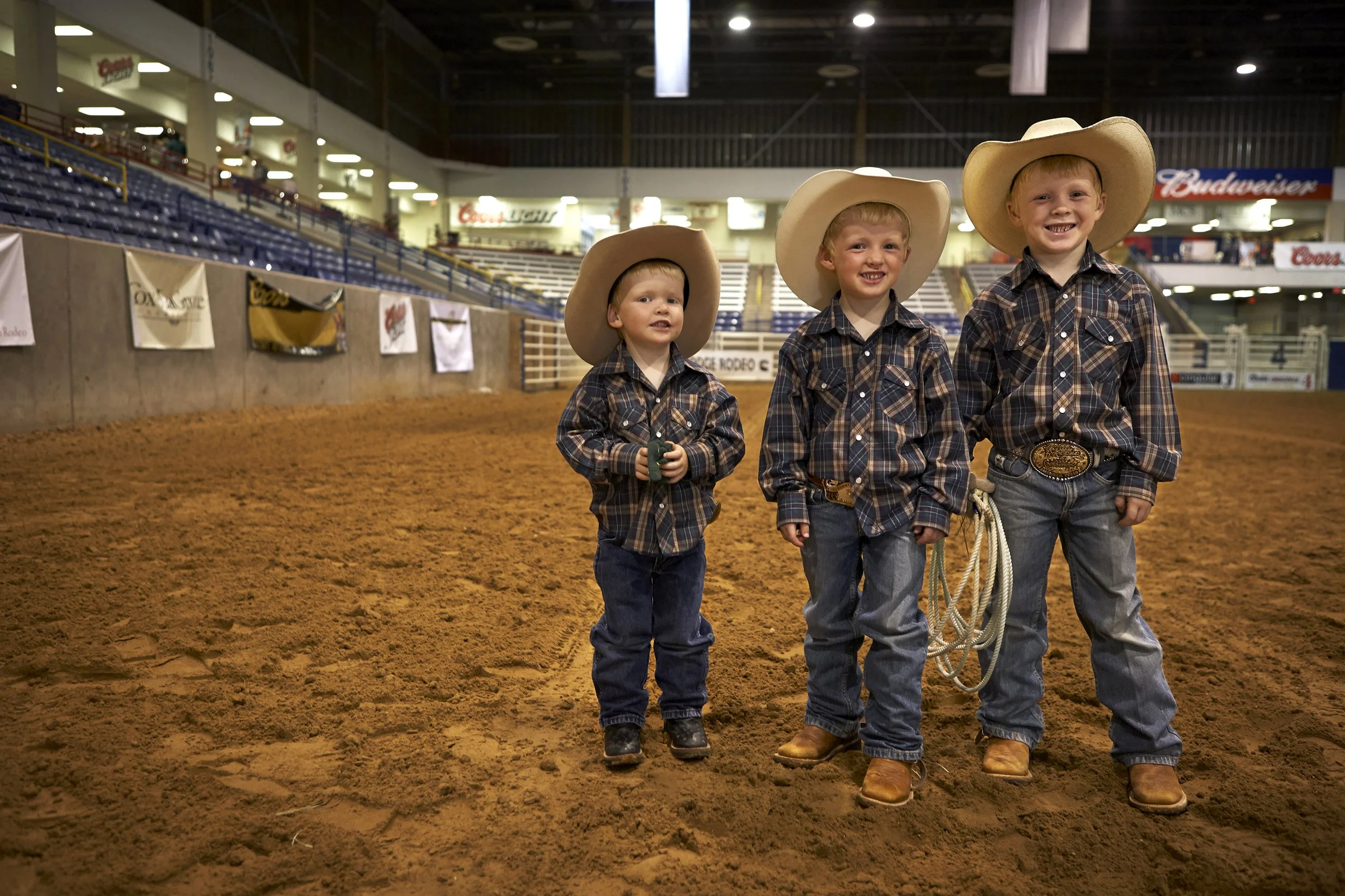 Three young boys wearing cowboy hats, plaid shirts, and jeans, standing on dirt inside an indoor rodeo arena with empty spectator seats and banners in the background. Andrew Fornasier Digital Tech San Francisco