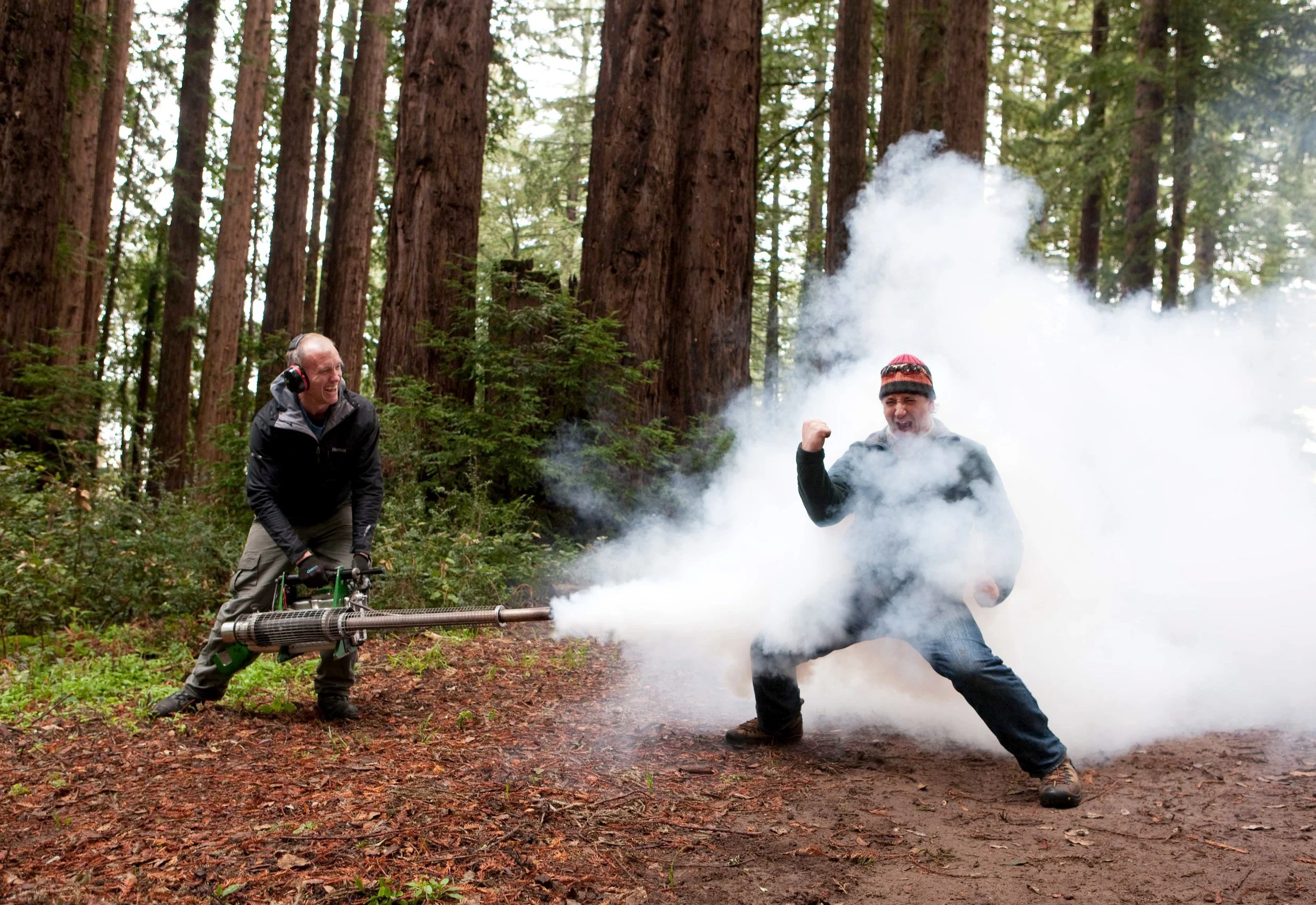 Two men in a forest, one wearing headphones and smiling, the other in a beanie, holding a device emitting smoke or vapor, with tall trees in the background.