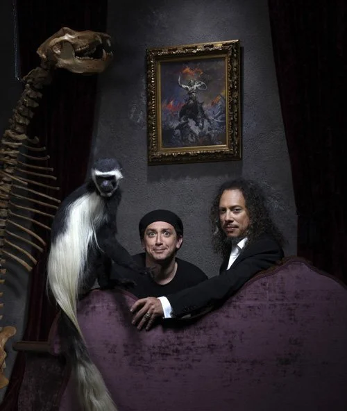 Two men sitting on a purple velvet sofa, with a monkey puppet, a dinosaur skeleton, and a framed painting of a stag on the wall behind them. Andrew Fornasier Digital Tech San Francisco