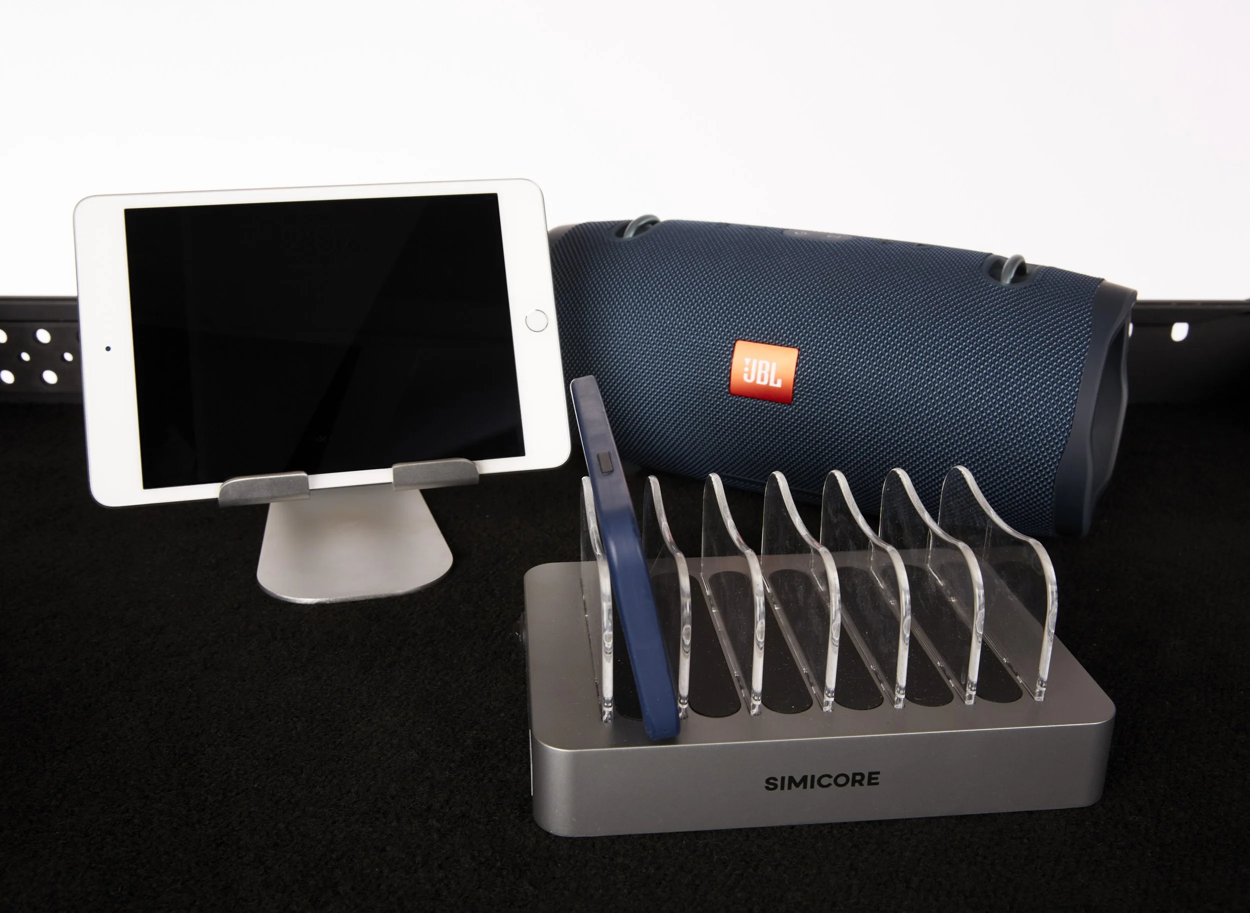 A white tablet on a stand, a blue portable speaker labeled JBL, and a set of utensils including a blue pen, all placed on a black surface and a silver dish rack with empty slots. Andrew Fornasier Digital Tech San Francisco