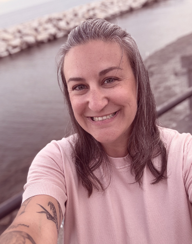 Woman with shoulder-length brown hair smiling and taking a selfie outdoors near a body of water with a rocky shoreline in the background.