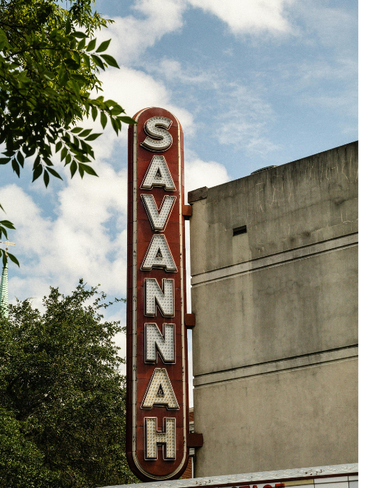 Vertical sign for Savanah with white illuminated letters on a red background, attached to an old concrete building, against a partly cloudy sky and some green tree leaves.