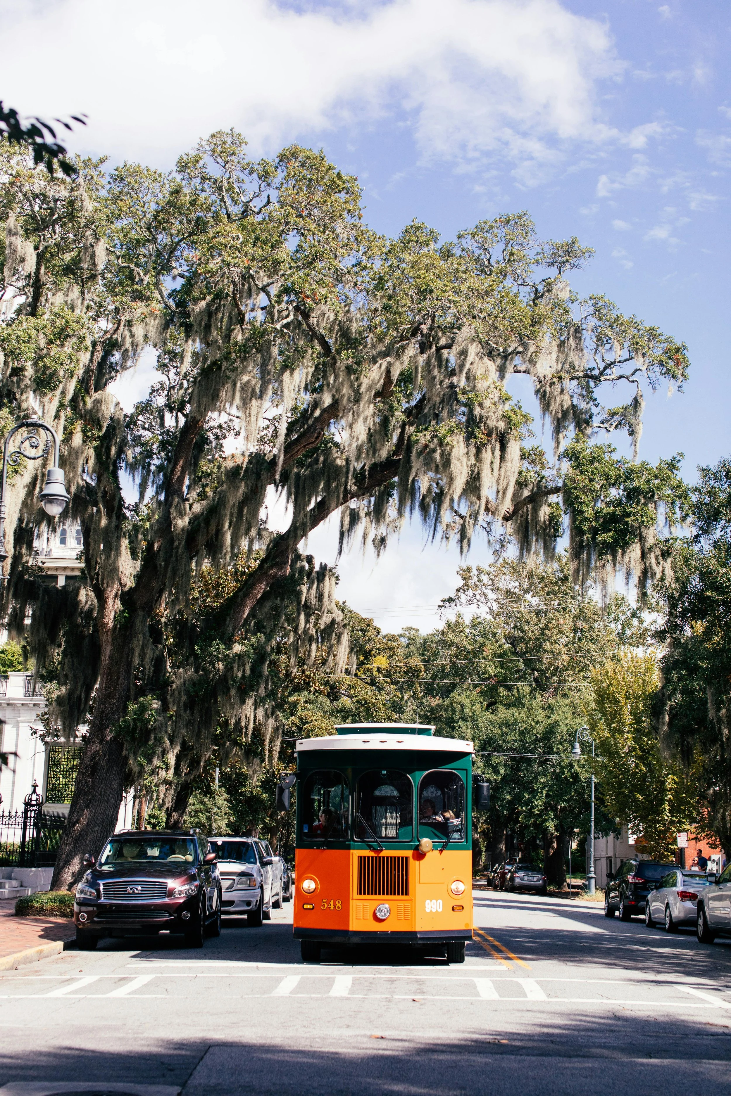 A vintage trolley bus painted orange and green traveling down a street with parked cars and tall trees with hanging moss.