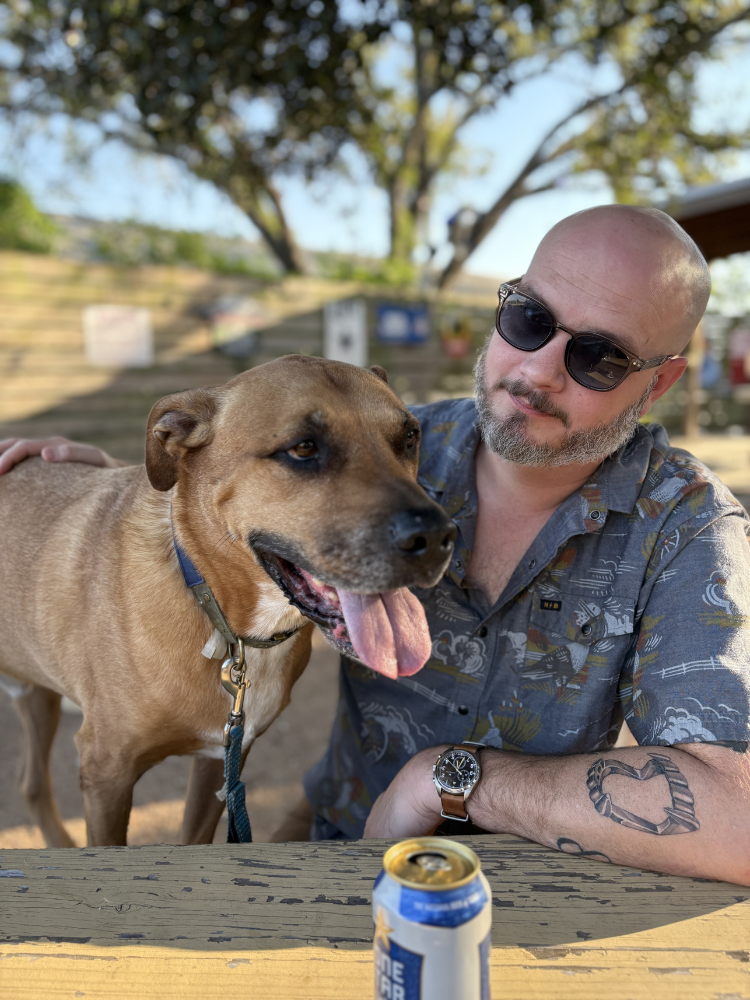 A man with sunglasses and a tattoo on his right forearm sitting at an outdoor table with a tan dog with a blue collar. There is a can of beer on the table and trees in the background.