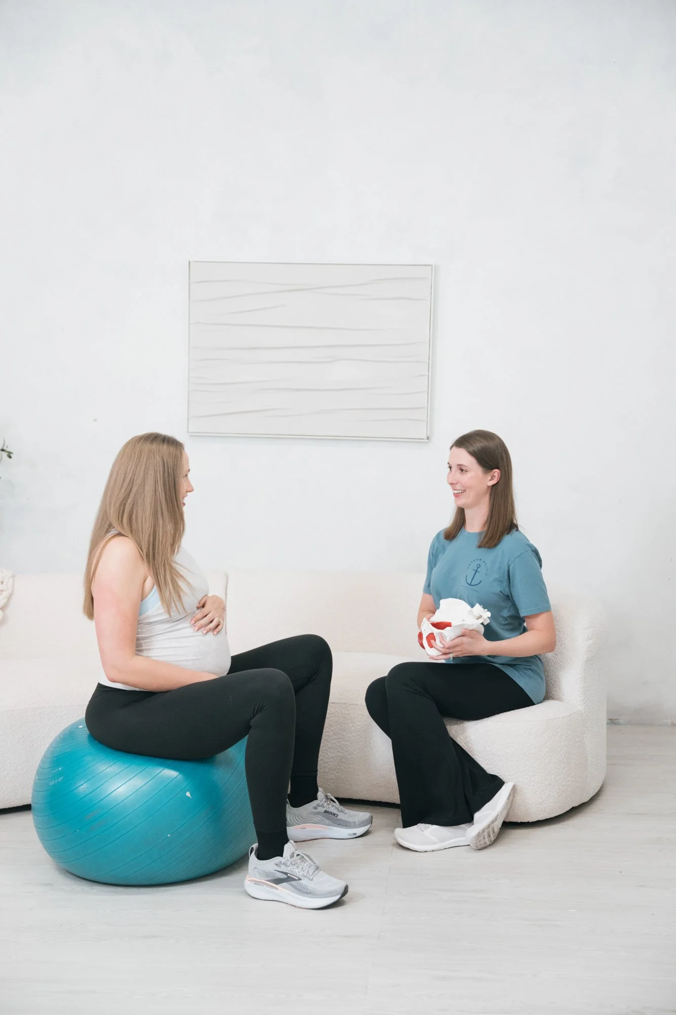 a pregnant woman sitting on a ball talking to a pelvic therapist who is holding a pelvis model