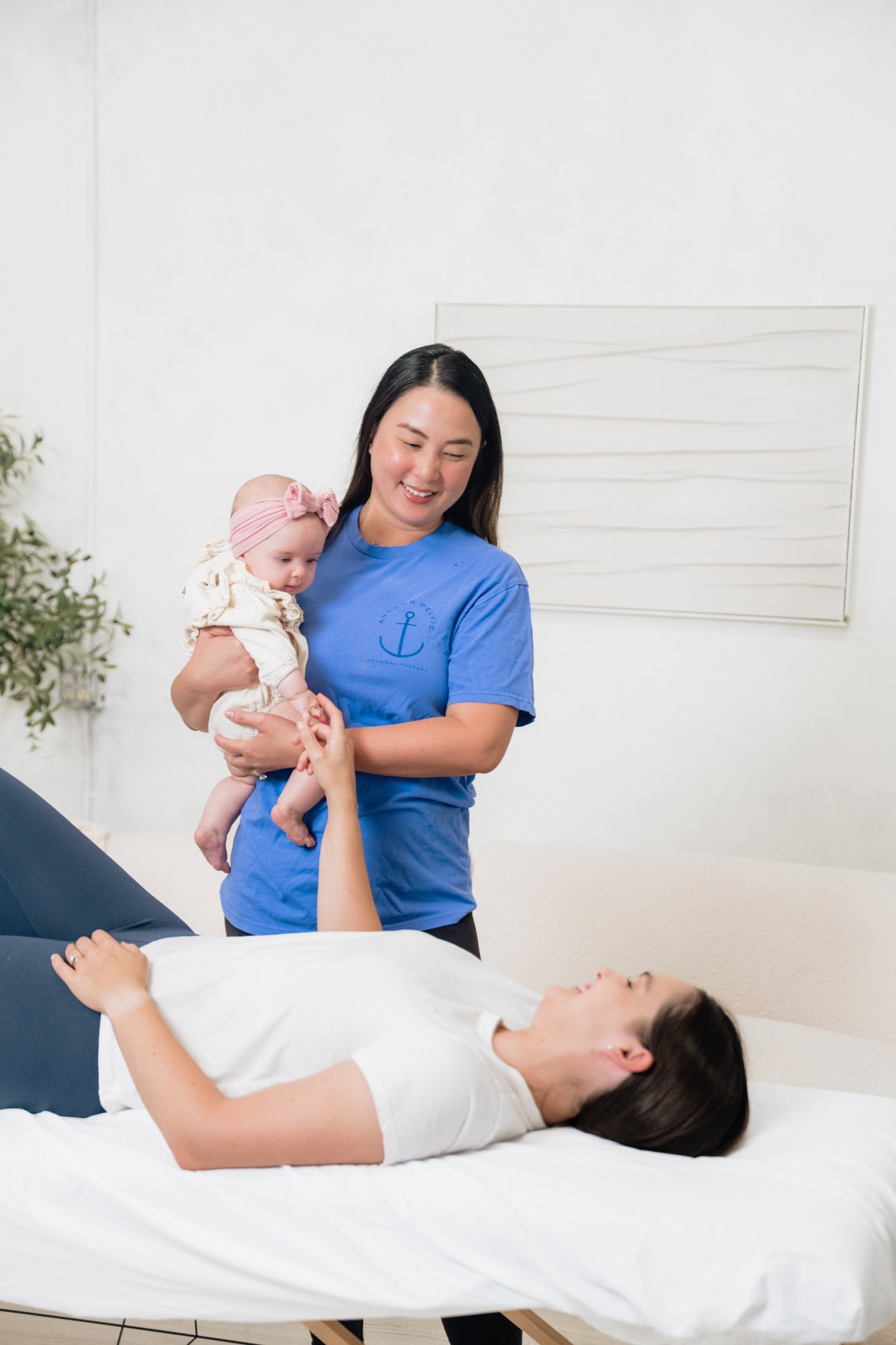A pelvic floor therapist holding a baby during a postpartum physical therapy session while the client lies on the table 