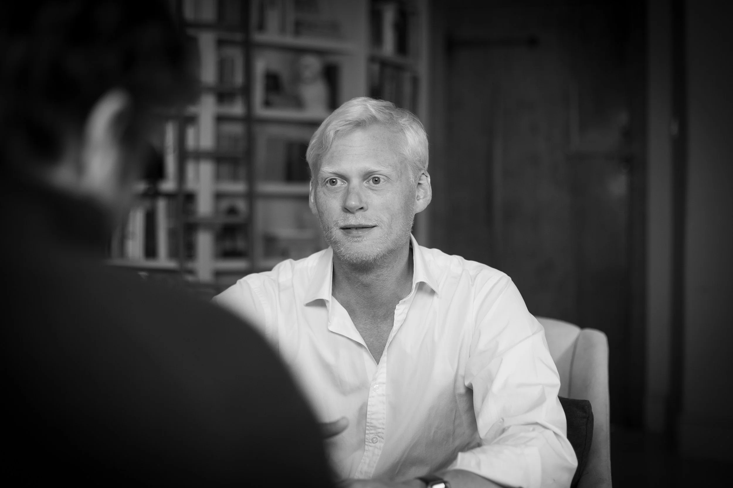 A black and white photo of a man with blond hair and a beard, sitting at a table and engaging in conversation with a woman who is partially visible in the foreground. The background features shelves filled with books.