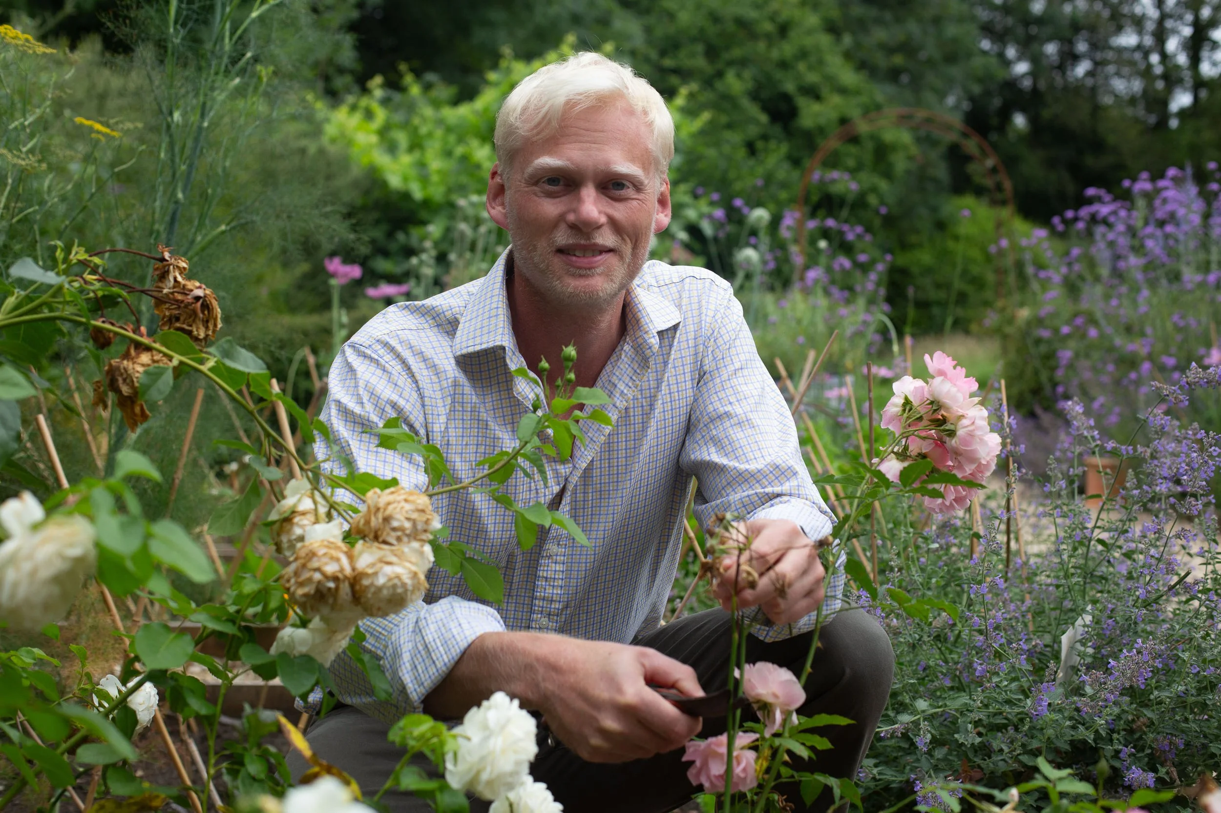 A man with blonde hair and a checked shirt kneeling in a lush garden surrounded by various blooming flowers, including roses and purple flowers, smiling at the camera.