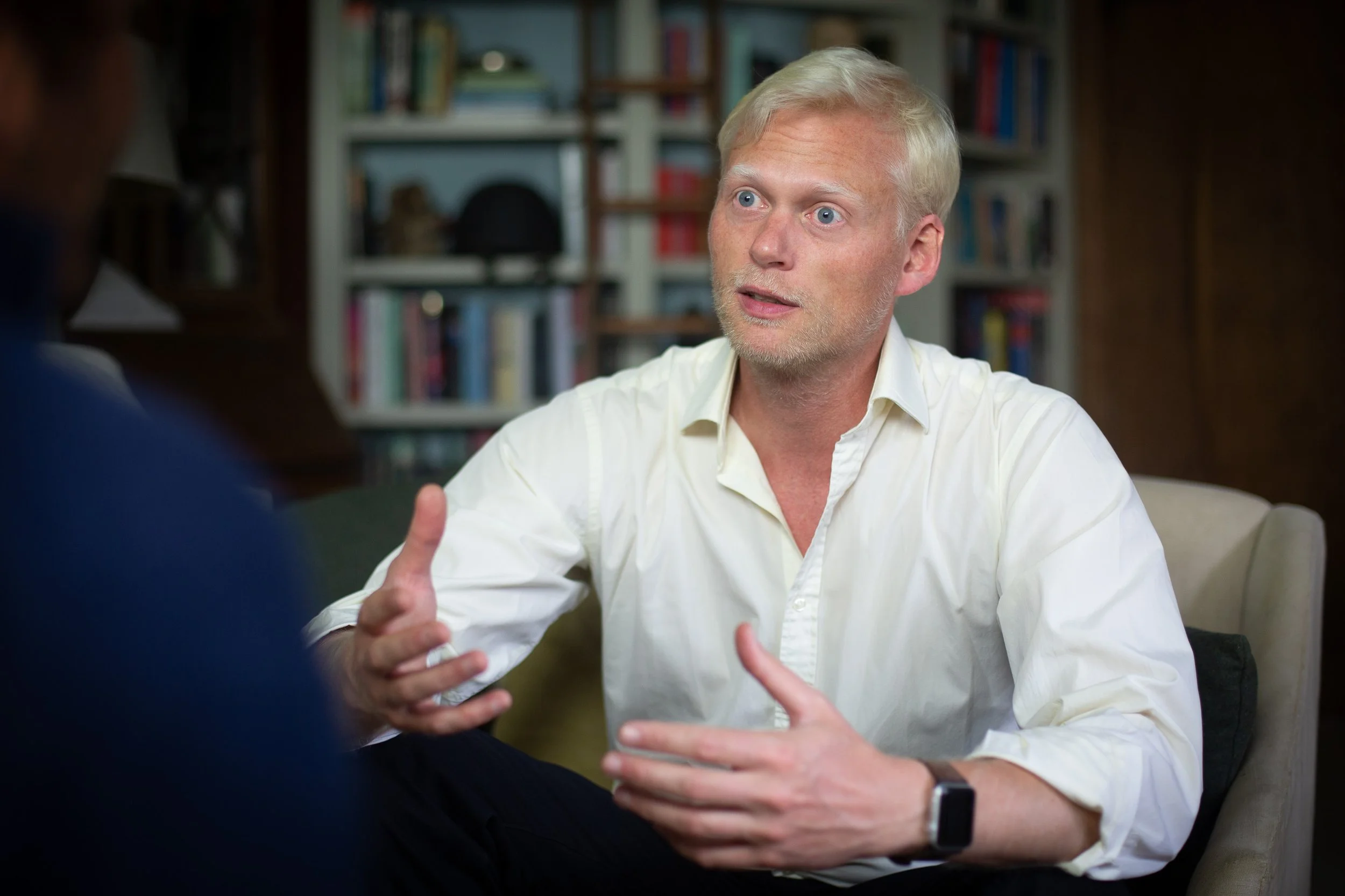 A man with blonde hair and a beard wearing a white shirt, engaged in a conversation with another person. He is gesturing with his hands and appears to be speaking. The background shows bookshelves with various books and decorative items.