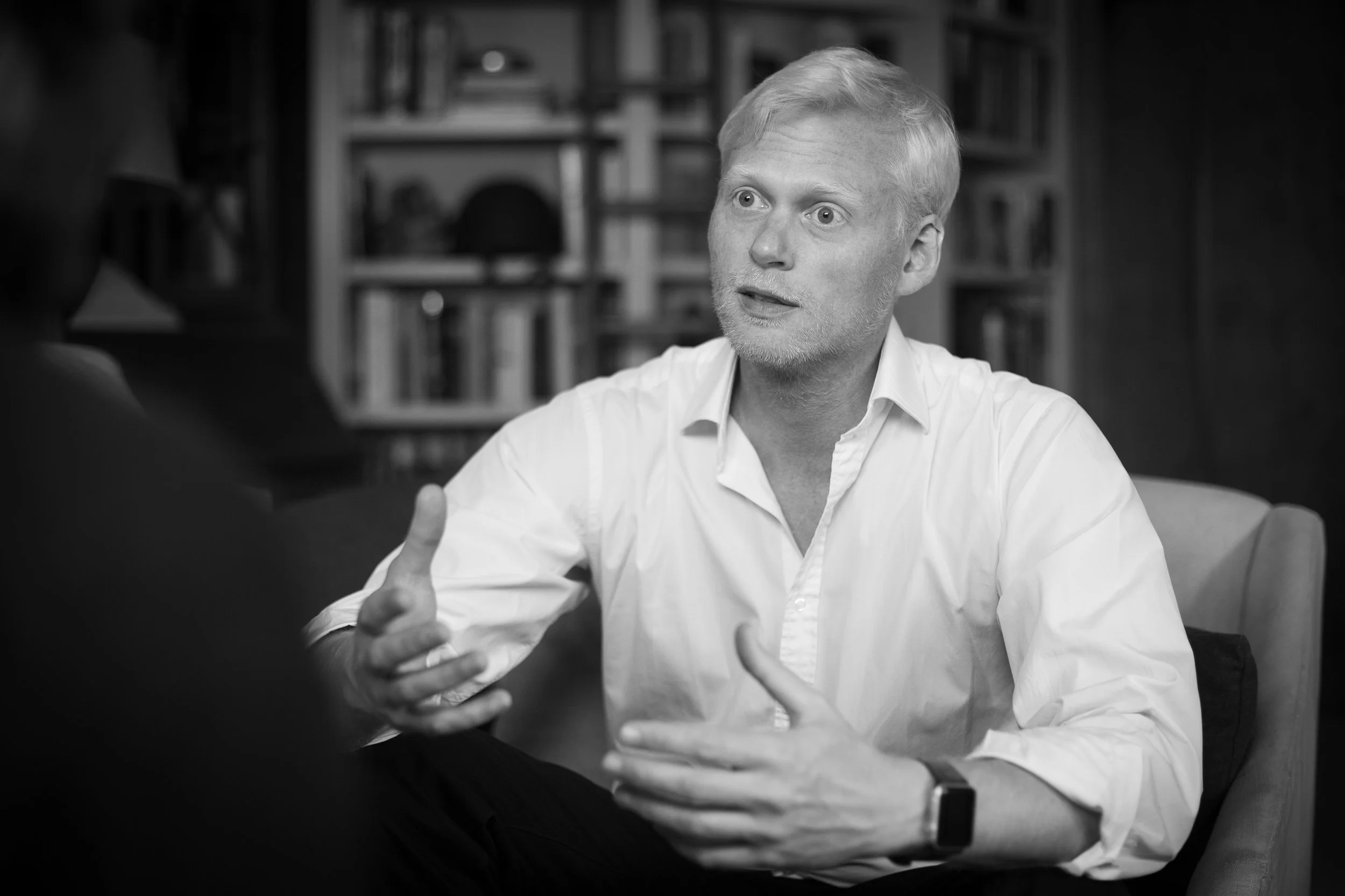 A black and white photo of a man with light hair and a beard, wearing a white shirt, engaged in a conversation, gesturing with his hand, in a setting with a bookshelf in the background.