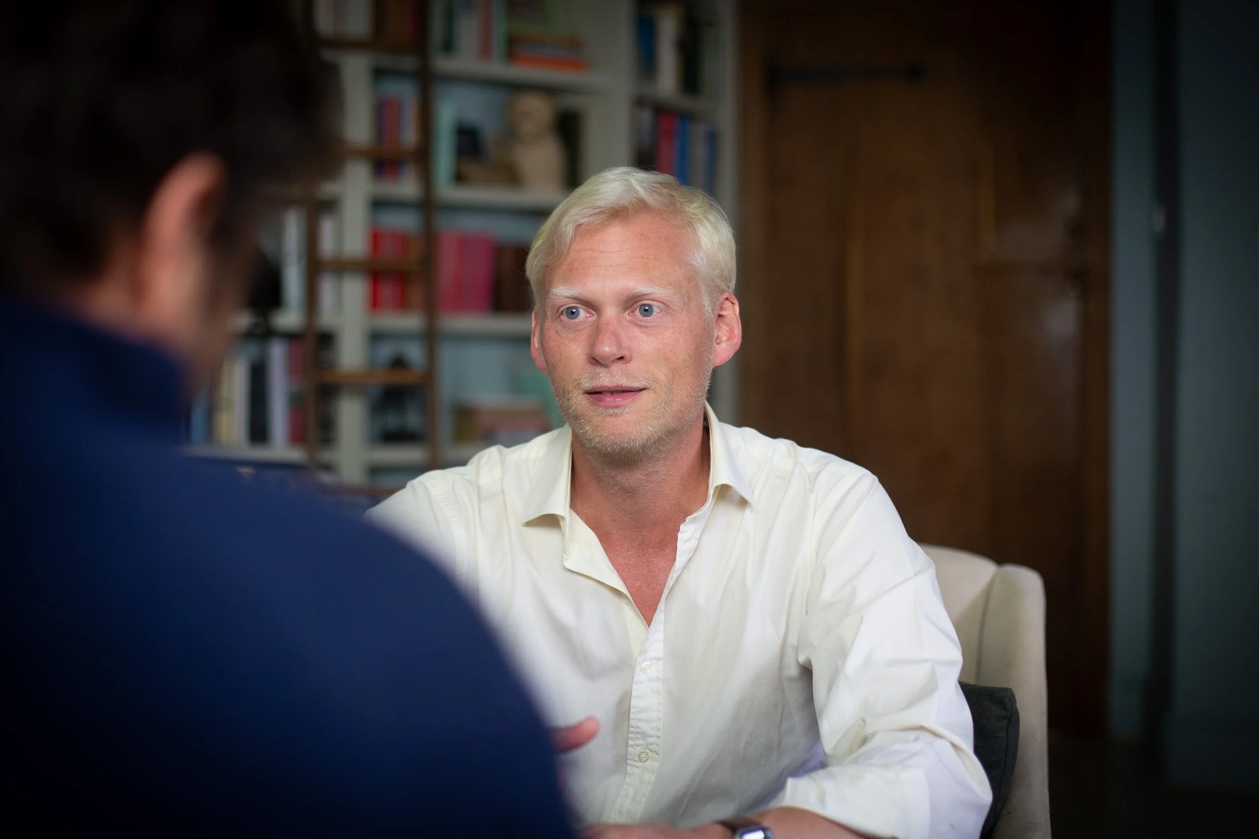 Two men are seated and engaged in a conversation. One man, with blonde hair and a light-colored shirt, is visible in the foreground, while the other person, with dark hair, is blurred and partially visible in the background. The setting appears to be a cozy room with bookshelves filled with books and decor in the background.