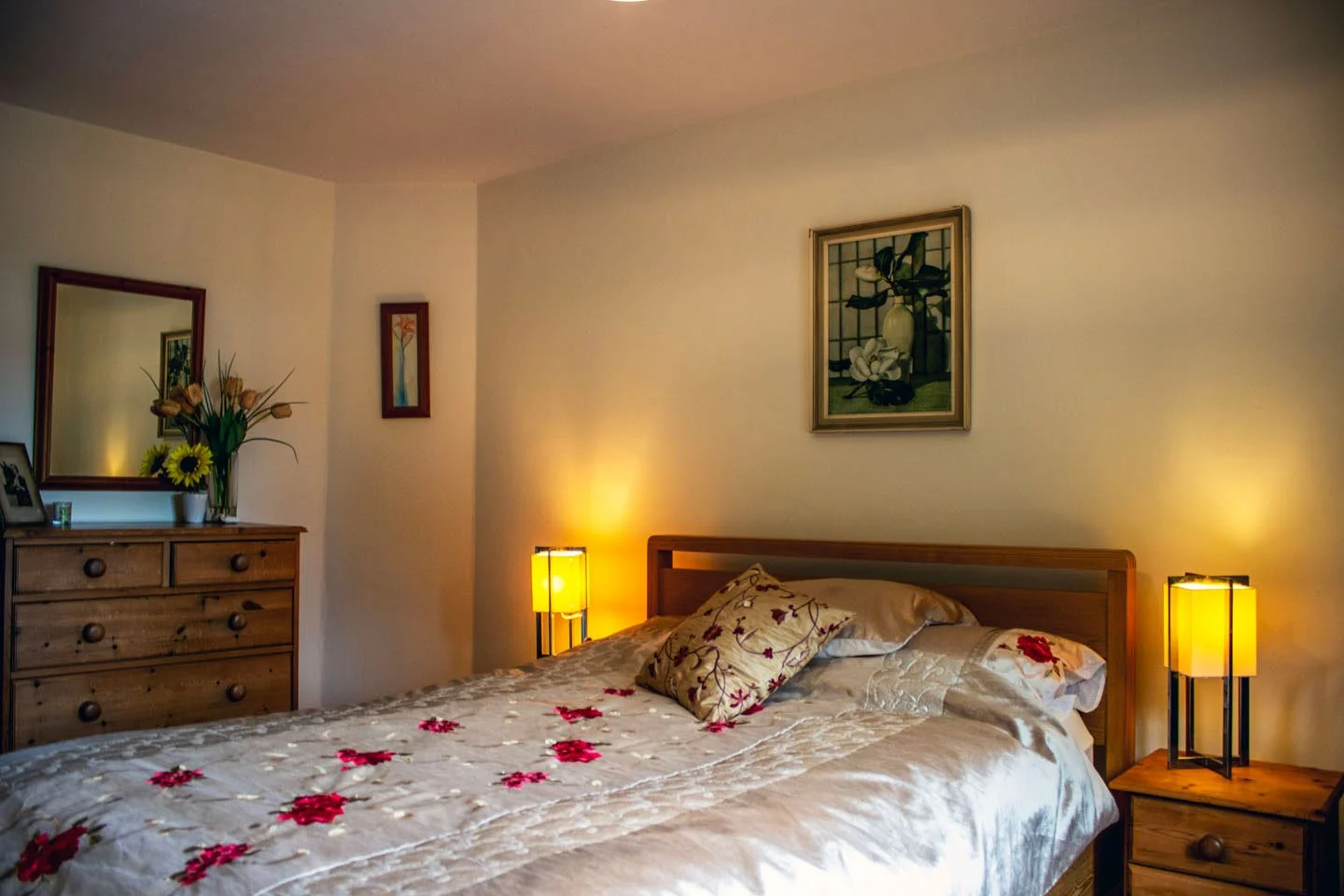 A cozy bedroom with a wooden bed, floral bedspread, and pillows. Two bedside tables with yellow lamps, a dresser with a mirror, flowers, and framed art on the walls.