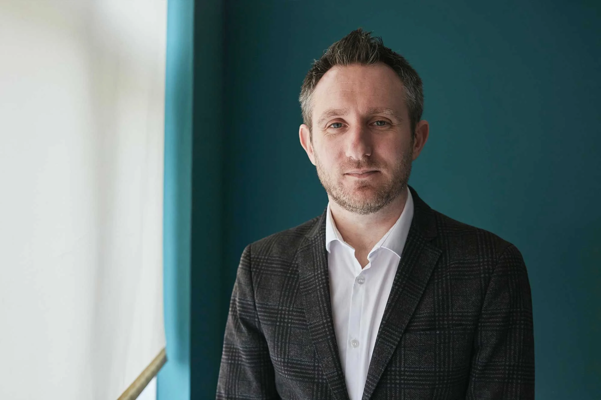 Portrait of a man with short dark hair, light stubble, wearing a white shirt and a dark checkered blazer, standing indoors near a window with a blue wall background.