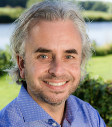 Close-up of a smiling man with gray hair and a beard, wearing a blue collared shirt, outdoors with a lake and green landscape in the background.