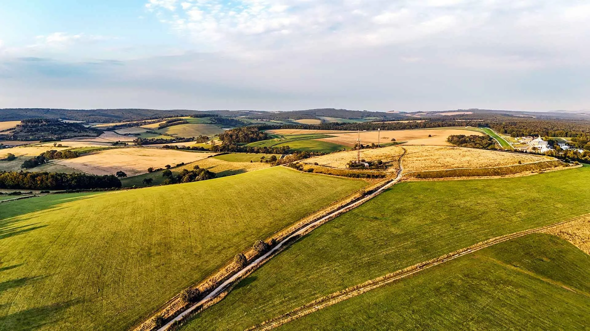 Aerial view of rolling farmland with green and yellow fields, dirt roads, trees, and distant hills under a partly cloudy sky.
