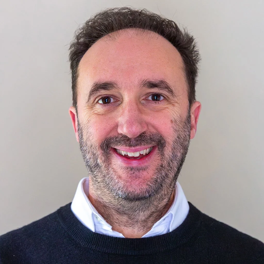 A smiling middle-aged man with dark hair and a beard, wearing a white collared shirt and a black sweater, standing against a plain background.