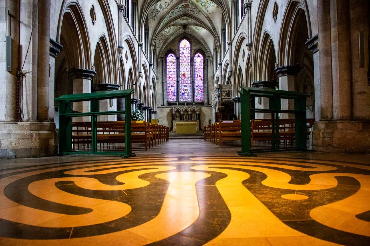 Interior of a church with stained glass windows, wooden pews, stone arches, and a patterned tile floor.
