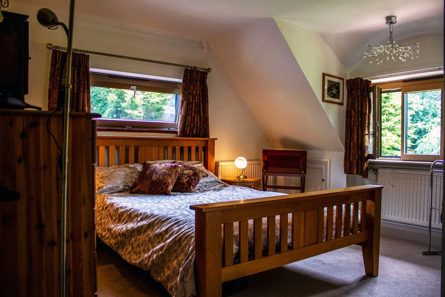 A cozy bedroom with a wooden bed, decorated with pillows, illuminated by a small bedside lamp, with windows showing green trees outside, carpeted floor, and a ceiling light fixture.