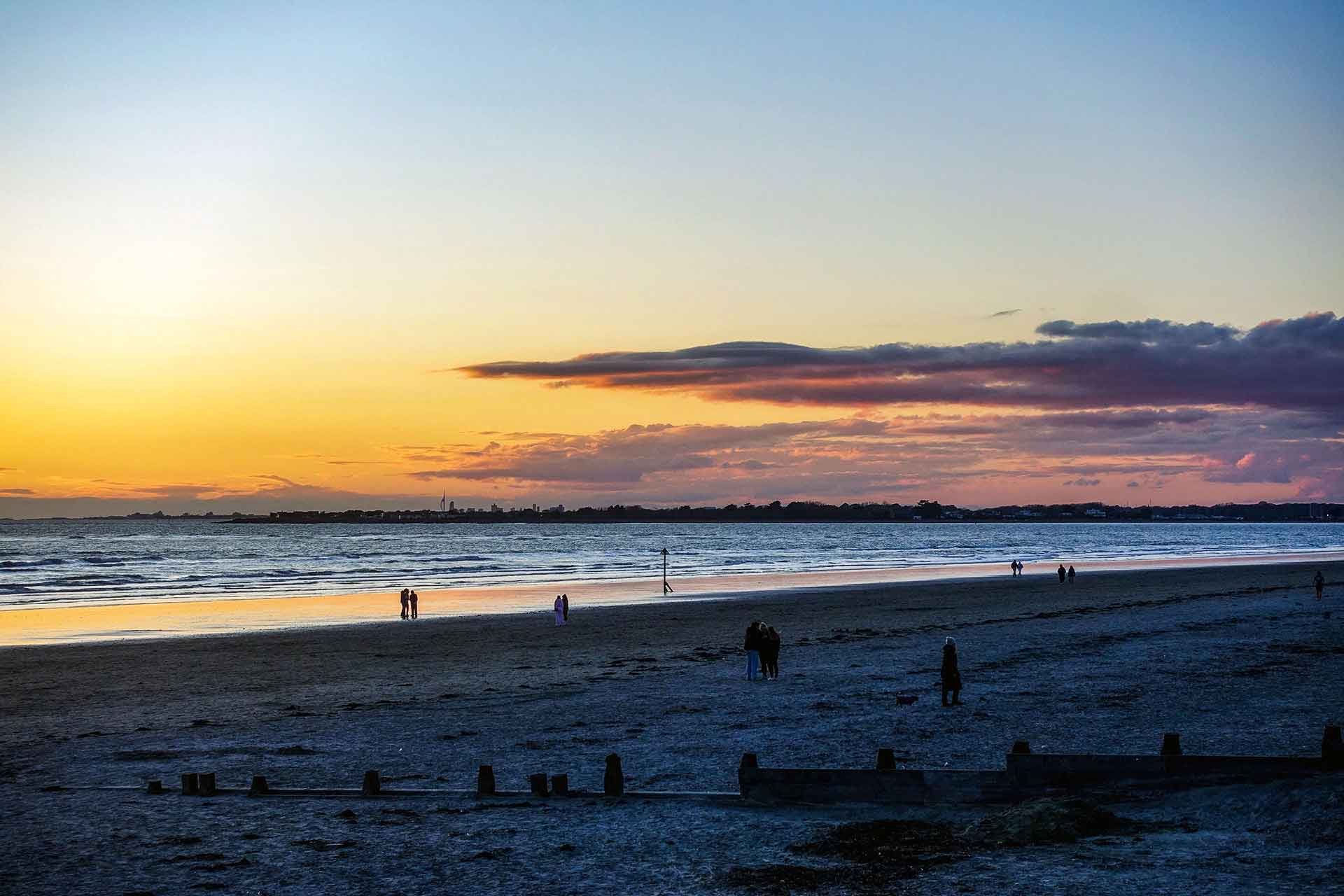 Sunset over the ocean with a partly cloudy sky, and people walking along the shoreline at the beach.
