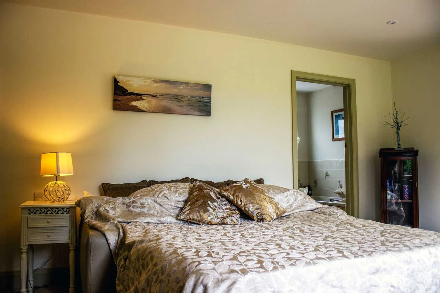 A cozy bedroom featuring a large bed with beige floral bedding and decorative pillows. A nightstand with a yellow lamp is on the left, and there is a painting of a beach scene hanging above the bed. A doorway leads to what appears to be a bathroom, and there is a wooden cabinet with a decorative tree on top on the right side of the room.